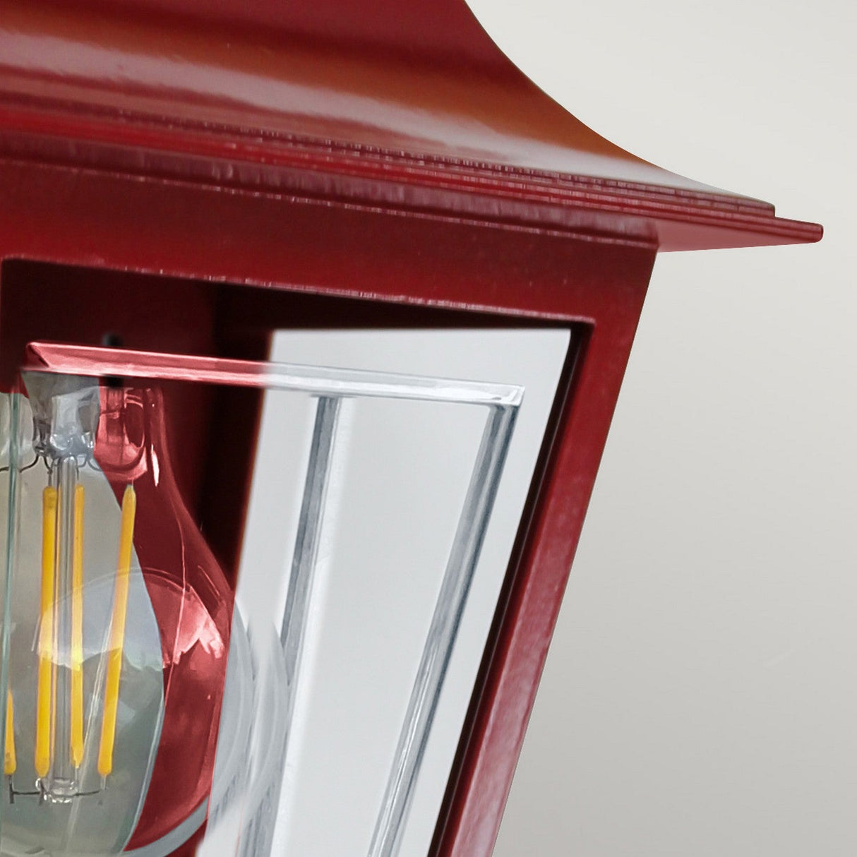 Close-up of the red Deco Outdoor Wall Lantern featuring a clear glass panel that reveals an LED bulb inside. The lantern, made from aluminum, is mounted against a light gray background.