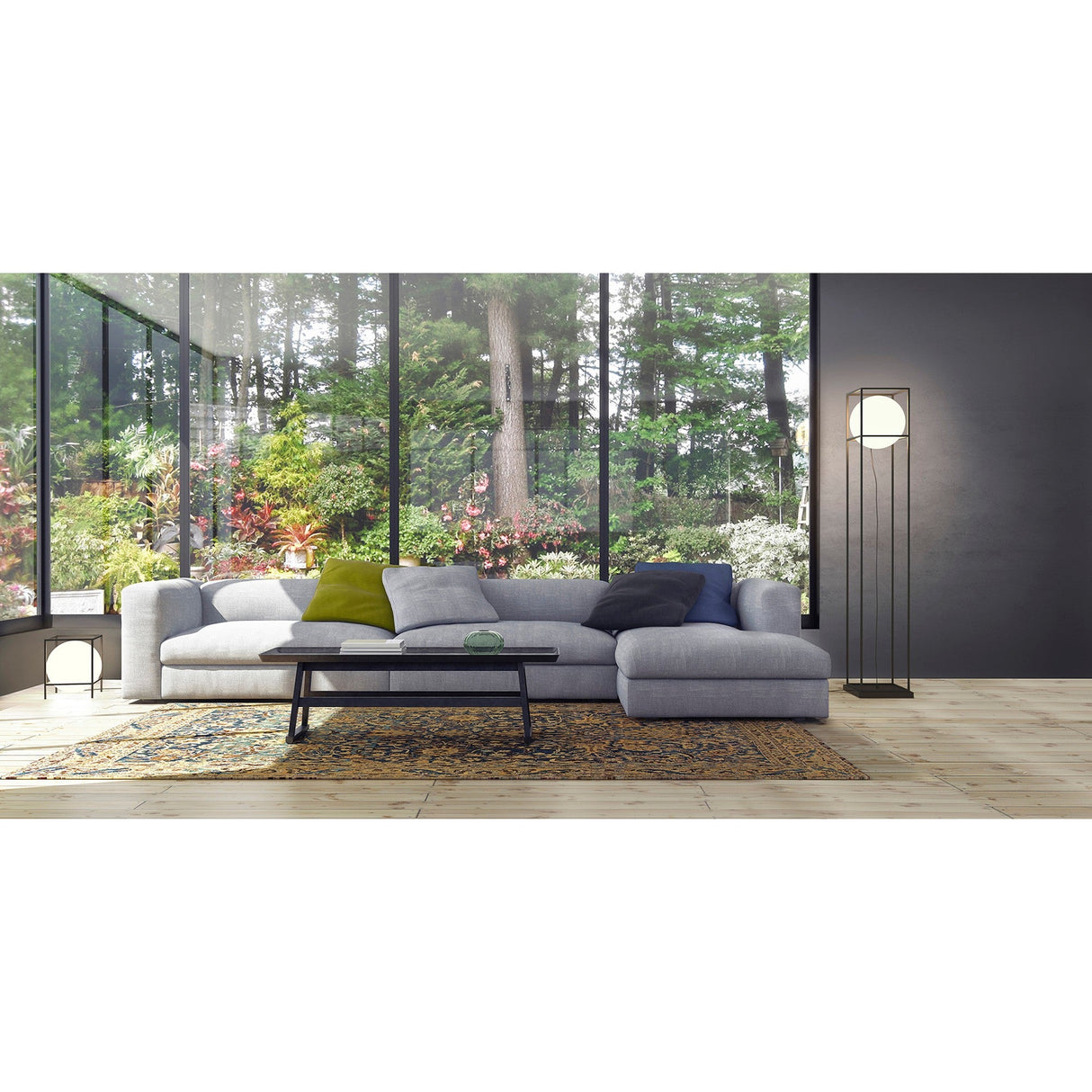A modern living room with a large gray sectional sofa, colorful pillows, a glass coffee table, patterned rug, the Desigual Large 1 Light Floor Lamp in matt black for ambient lighting, and large windows revealing a lush green garden.