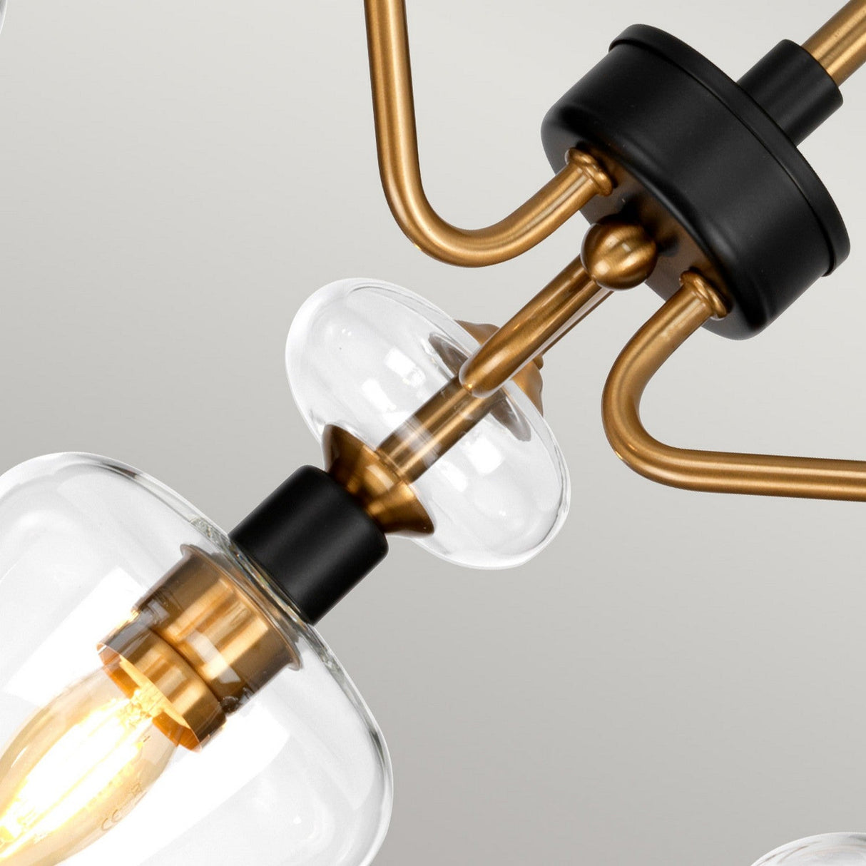 A close-up of the Armand 3 Light Pendant Semi-Flush reveals multiple light bulbs enclosed in clear glass, accompanied by gracefully curved arms. The fixture features aged brass plated and charcoal black painted accents, highlighting the warm glow from a lit bulb.