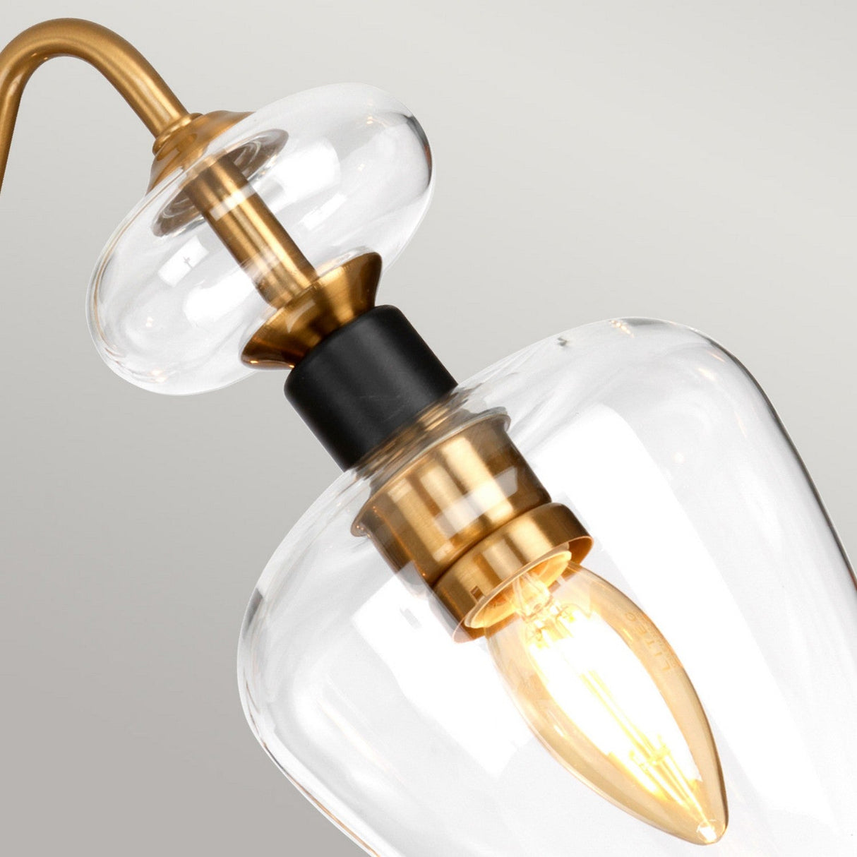 A close-up of the Armand 3 Light Pendant Semi-Flush - Aged Brass Plated & Charcoal Black Paint highlights its modern design with a clear glass shade and accents of aged brass and charcoal black paint. Inside, a glowing bulb casts a warm light against the soft, light gray background.