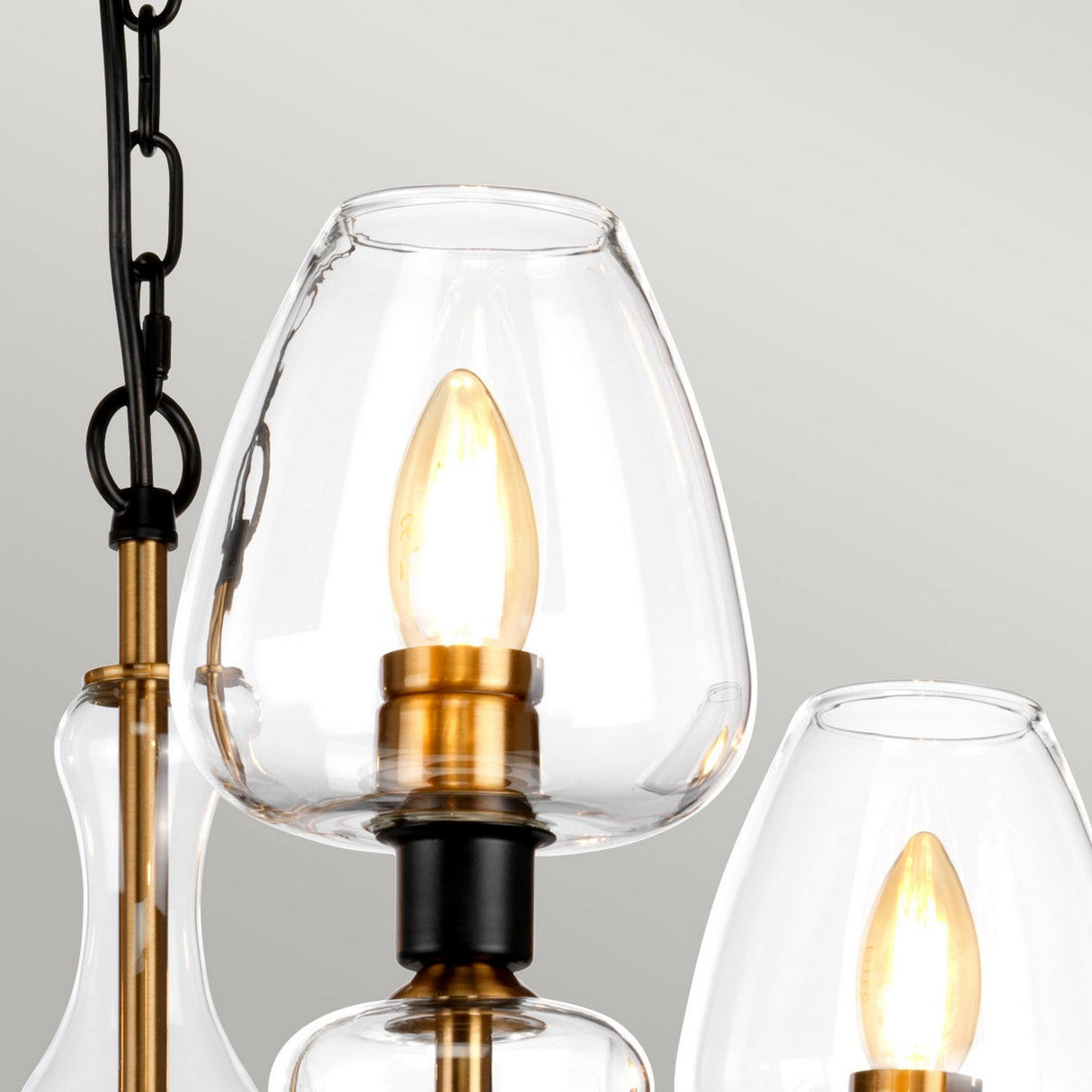 Close-up of the Armand 3 Light Chandelier, showcasing its contemporary design with clear glass shades and exposed bulbs. The metal fixtures are finished in aged brass plating and charcoal black paint, embodying modern lighting design, and emit a warm glow. A black chain is partially visible on the left.