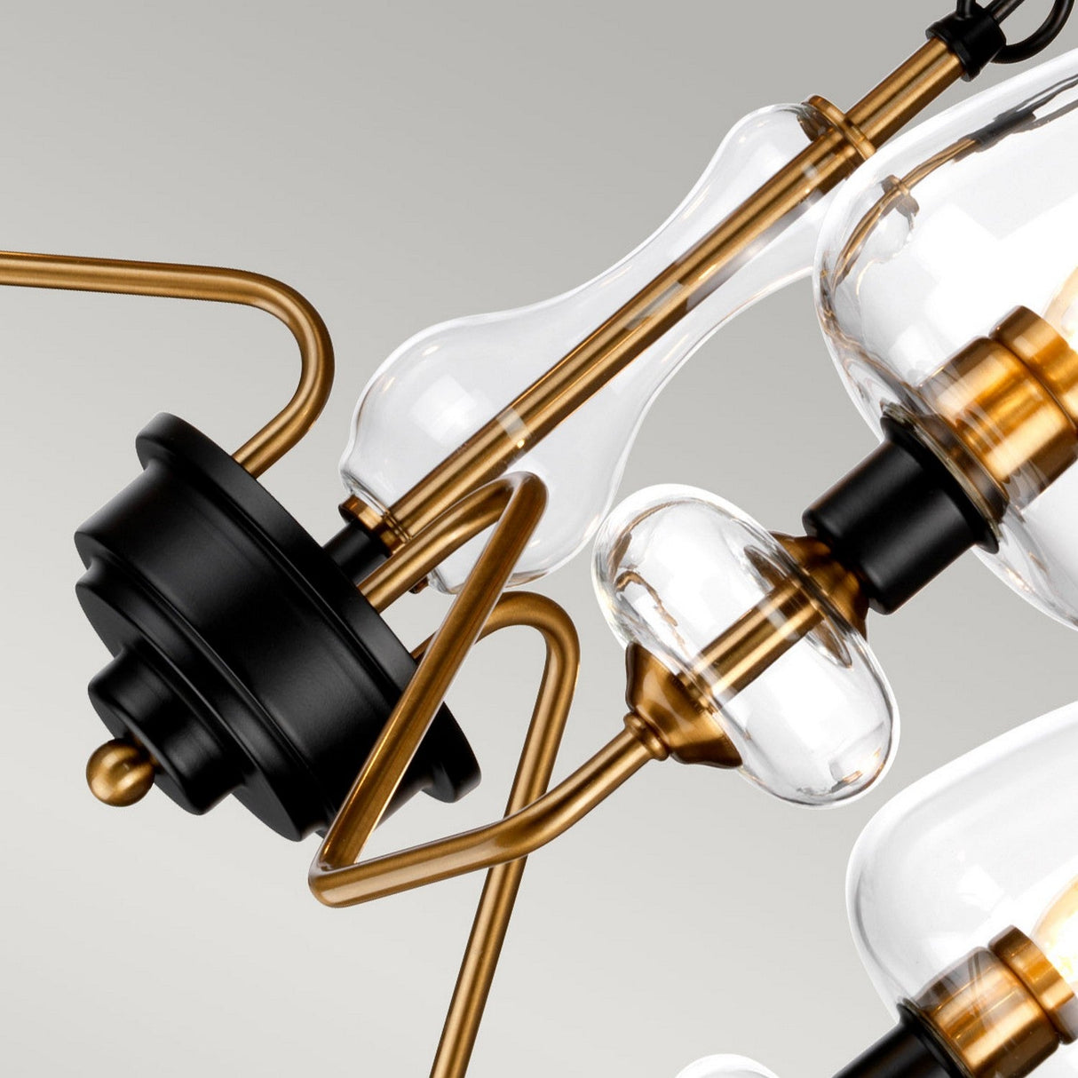 Close-up of the Armand 3 Light Chandelier highlighting its clear glass globes and stylish aged brass plated components, complemented by charcoal black paint. This modern lighting design harmoniously combines curved and straight lines set against a light gray backdrop.