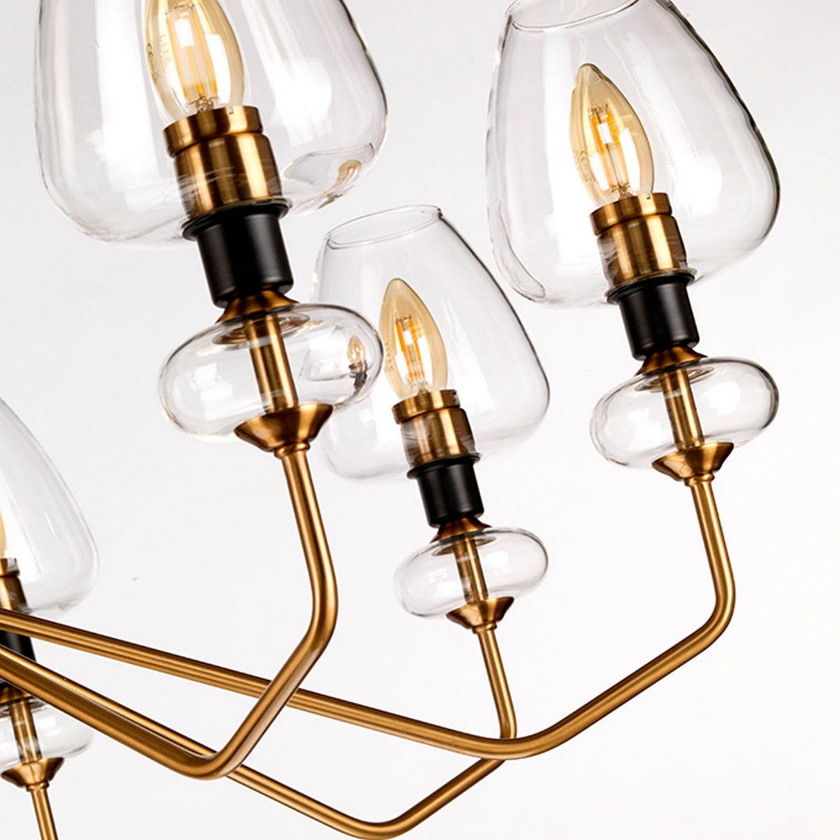 Close-up of the Armand 8 Light Chandelier, highlighting its aged brass plated metal arms and charcoal black paint accents with clear glass shades. It features exposed bulb filaments and elegantly curved structures, all presented in a sleek, contemporary design against a pristine white background.