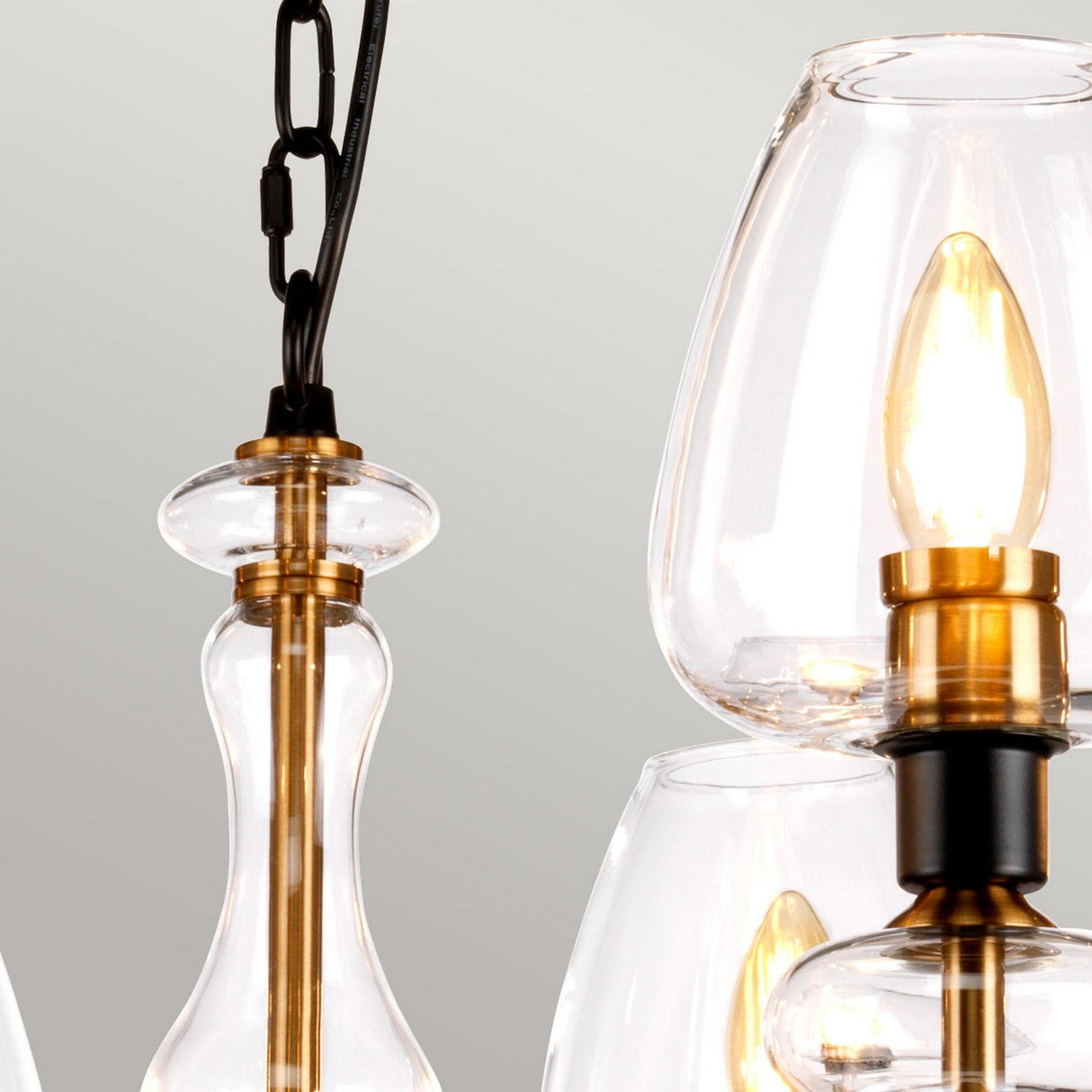 Close-up of the Armand 8 Light Chandelier, showcasing its contemporary design with clear glass lampshades and aged brass plated fittings accented by charcoal black paint. The image highlights the intricate design and visible bulbs inside. The chain and wire extend from the fixture, standing out against a light gray background.