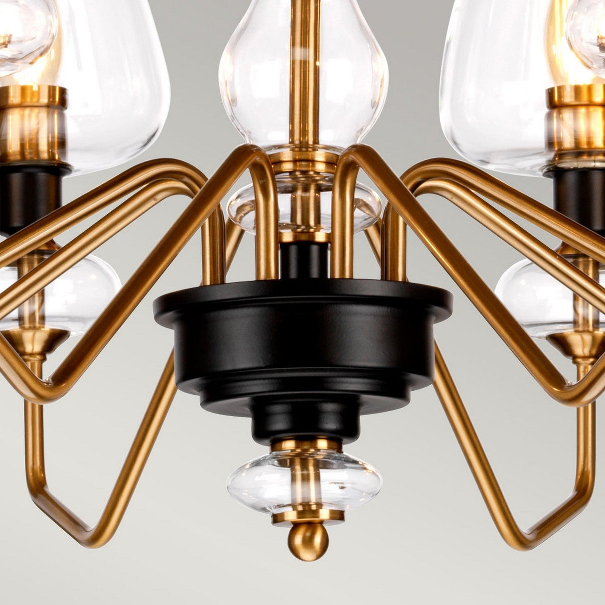 Close-up of the Armand 8 Light Chandelier, showcasing its aged brass plated and charcoal black painted finish. The contemporary design incorporates clear glass elements and multiple symmetrically extending curved arms against a neutral gray background.