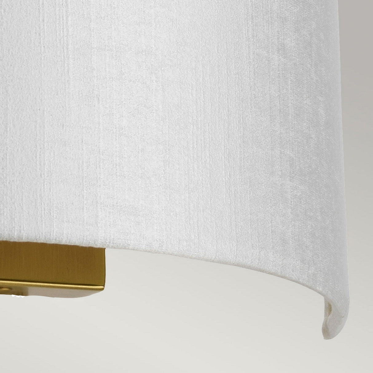 A close-up view of the Cooper Medium Curved Wall Light in aged brass showcases its white faux silk shade with a subtle texture, set against a light gray background.