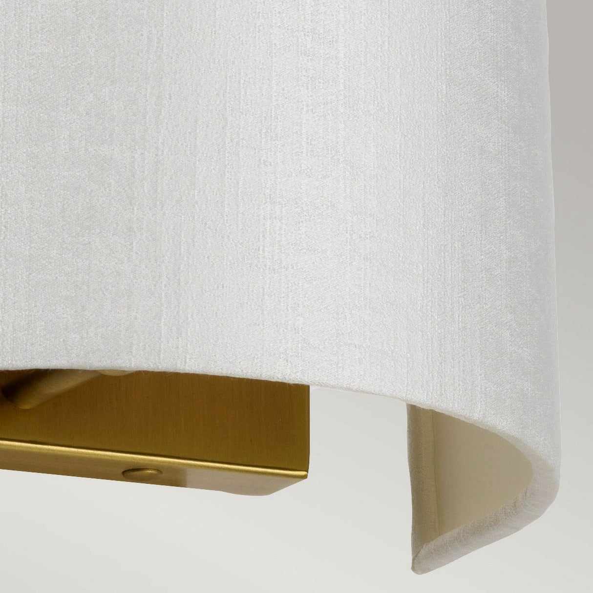 Close-up of the Cooper Small Curved Wall Light in aged brass, featuring a white textured fabric lampshade with an elegantly curved bottom edge. The visible mount in gold metal offers a sleek, modern element, ideal for ambient lighting.