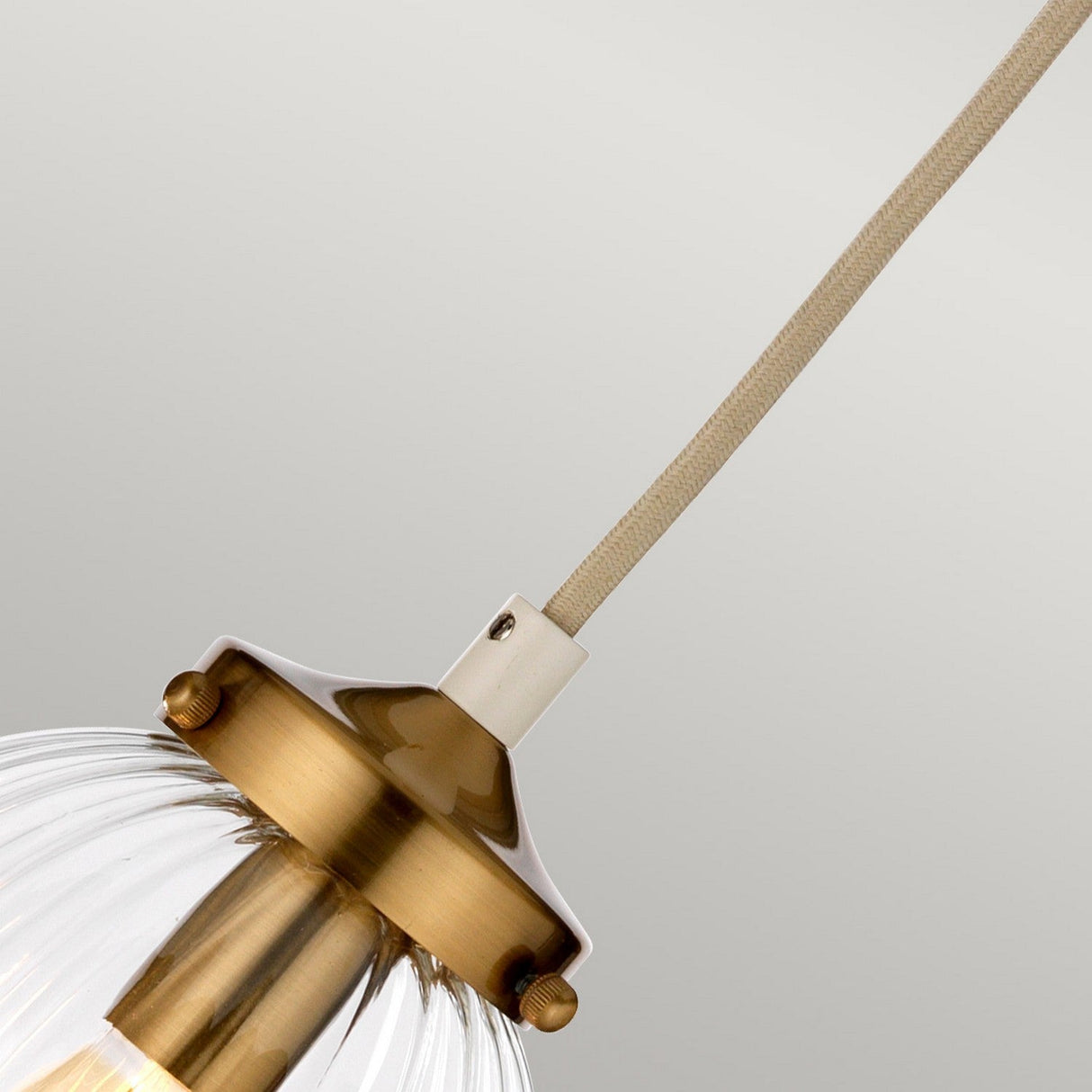 A close-up of the Cosmos 1 Light Pendant showcases its aged brass accents and clear hand-blown glass shade. It hangs from a cream-painted fabric-covered cord, set against a neutral background, offering a hint of retro style.