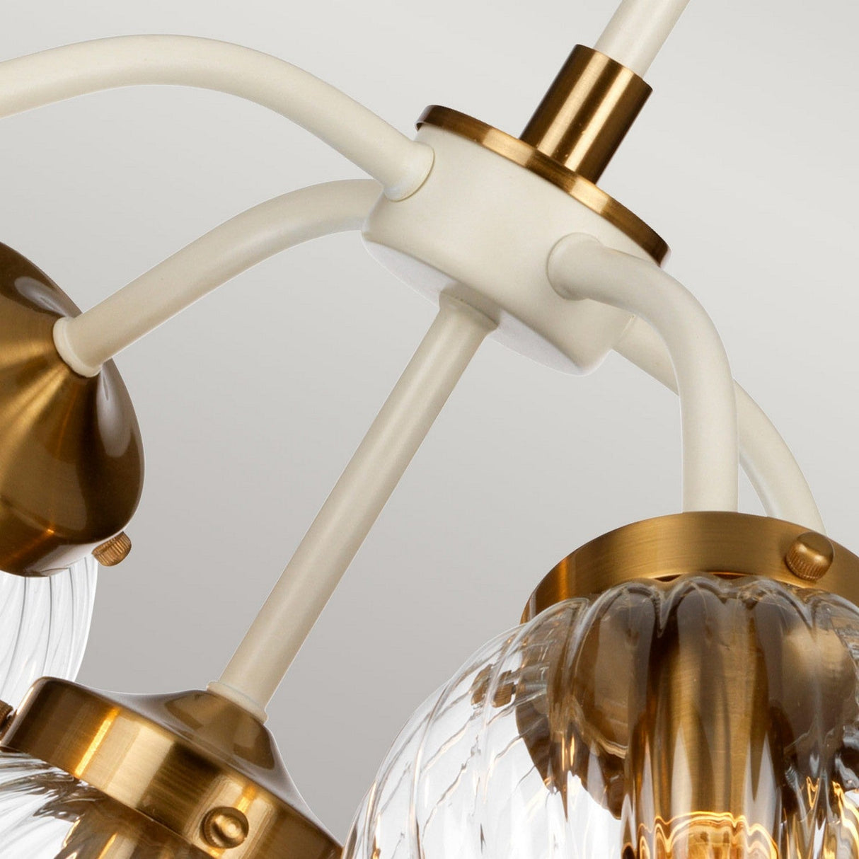 A close-up of a modern ceiling light fixture showcases three arms, each supporting a bulb within a fluted, hand-blown glass shade. The combination of white and brass offers an elegant, retro-inspired design that evokes the look of the Cosmos 5 Light Pendant in Cream Painted & Aged Brass against a neutral backdrop.