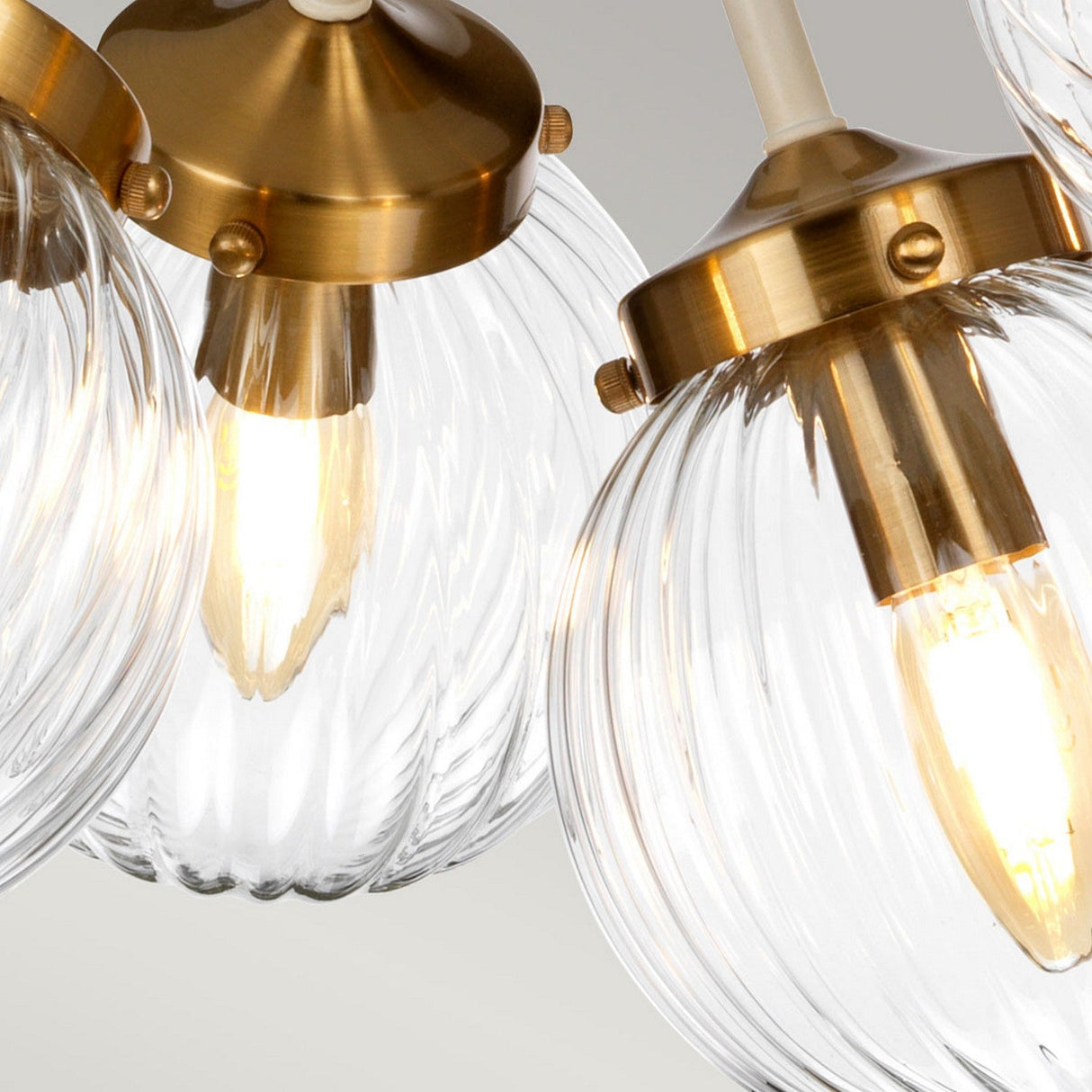 Close-up of three clear glass pendant lights featuring hand-blown, ribbed spherical shades and warm glowing bulbs. These fixtures, from the Cosmos 5 Light Pendant - Cream Painted & Aged Brass collection, are accented with brass hardware, emphasizing their retro-inspired modern design against a light gray background.
