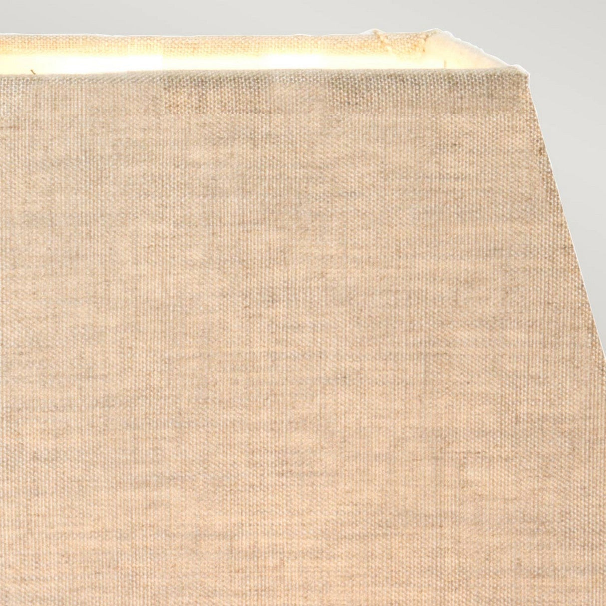 Close-up of a beige, textured fabric lampshade with visible light emanating from the top right, set against a pale gray background. Echoing the Eclipse Table Lamp With Tapered Square Shade - Bronze Patina design, the fabric looks worn, showing some fraying at the edges.