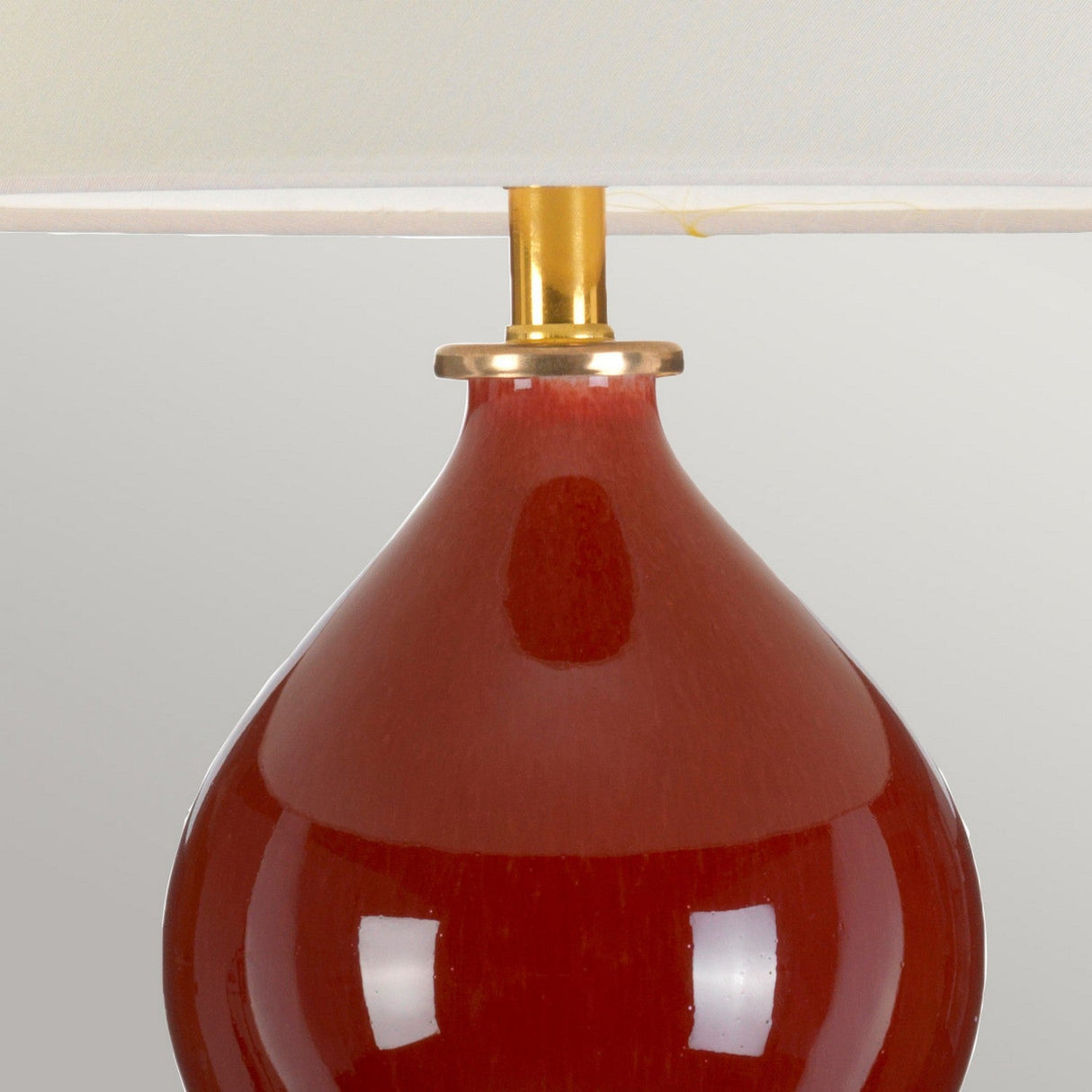 Close-up of the Harbin Gourd Table Lamp With Tall Empire Shade - Oxblood, showcasing its oxblood ceramic base with a shiny finish. The rounded shape is complemented by a gold-colored trim connecting the base to the tall empire shade, all set against a neutral background.