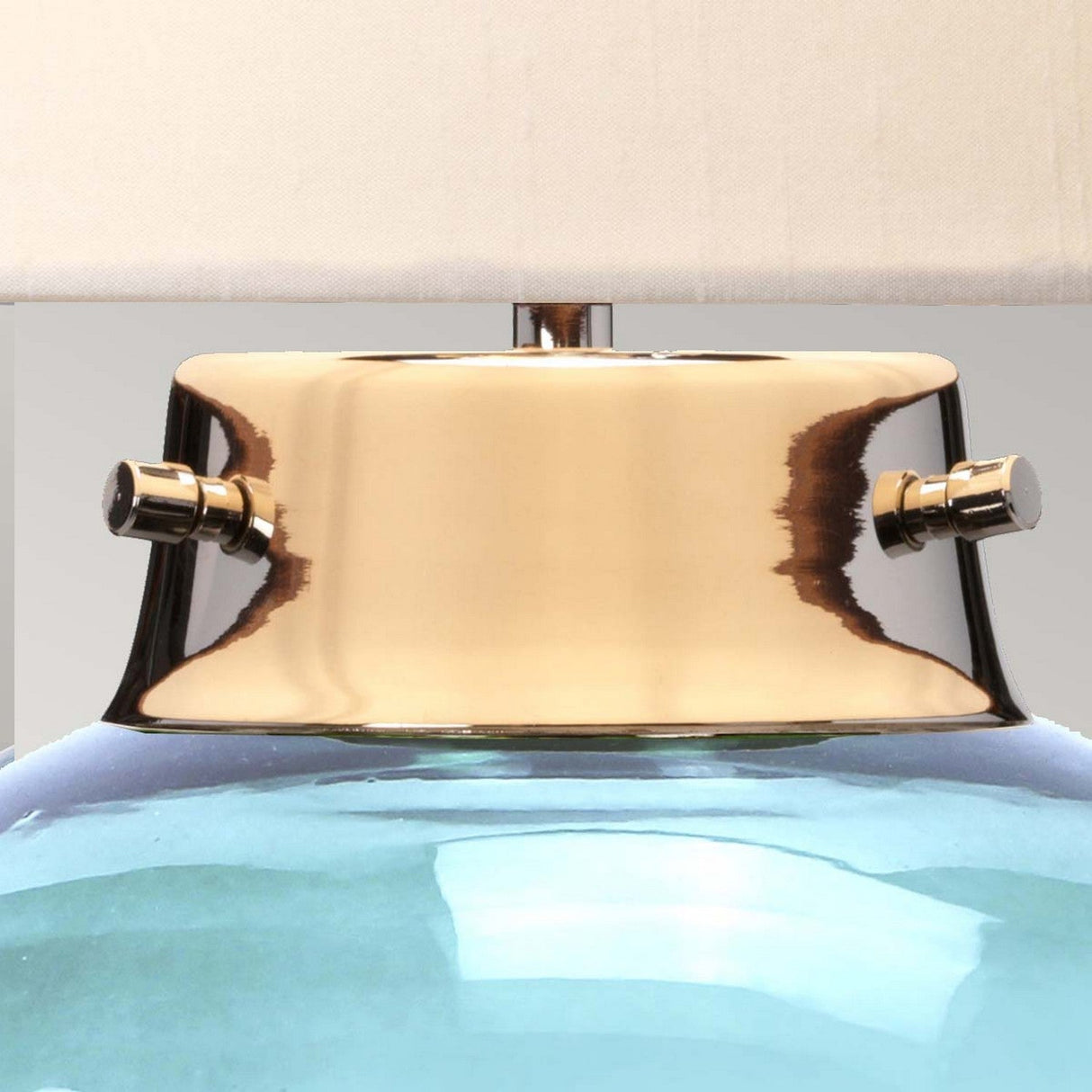 Close-up of the Kara Table Lamp's metallic neck in polished nickel, featuring two adjustment knobs with a reflective gold-like finish. Below is its vibrant blue glass base, and above, a portion of the white lampshade. The background is a neutral light gray.