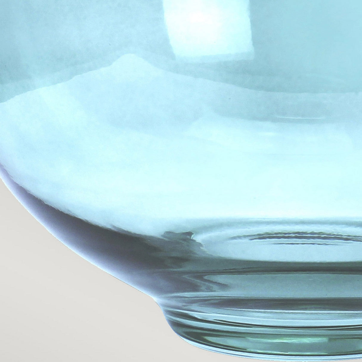 A close-up of a translucent, light blue glass bowl against a plain background mirrors the sophisticated aesthetic of the Kara Table Lamp - Polished Nickel - Blue Glass. The bowl's gentle curves and smooth surface catch and reflect the light, creating delicate highlights and gradients, similar to the lamp's polished nickel accents. Its visible lower edge adds to its charm.