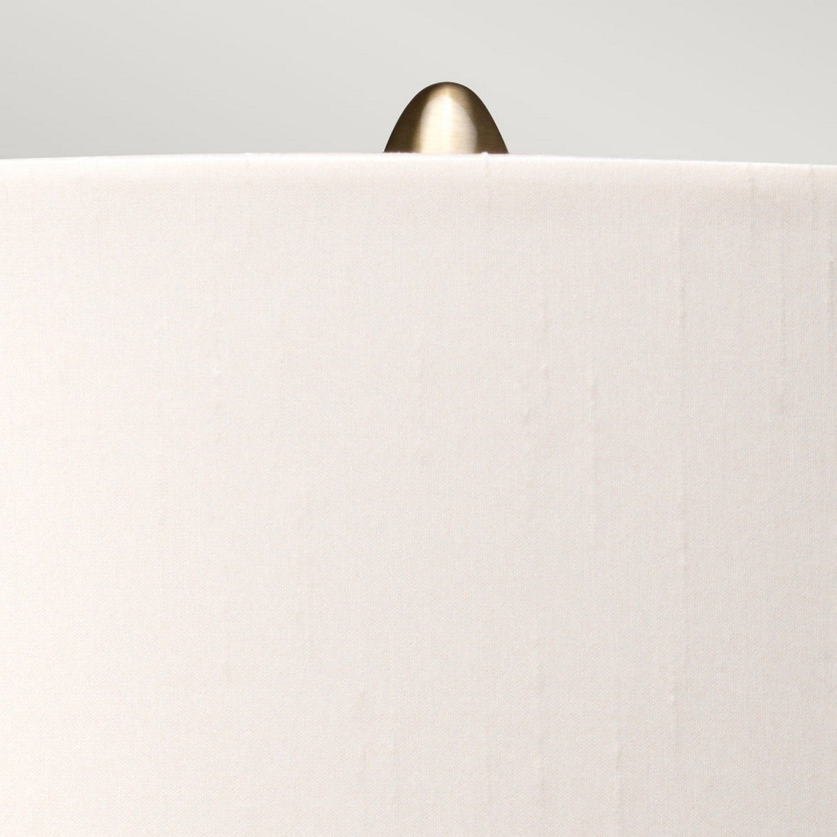 Close-up of a beige fabric lampshade with an aged brass finial on top, featured in the sophisticated Kara Table Lamp - Aged Brass - Dark Green Glass collection, set against a light gray background.