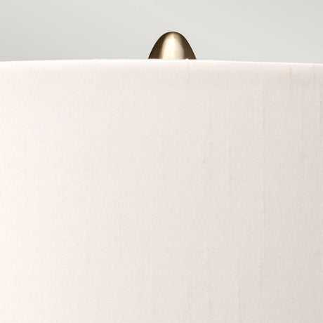 Close-up of a beige fabric lampshade with an aged brass finial on top, featured in the sophisticated Kara Table Lamp - Aged Brass - Dark Green Glass collection, set against a light gray background.