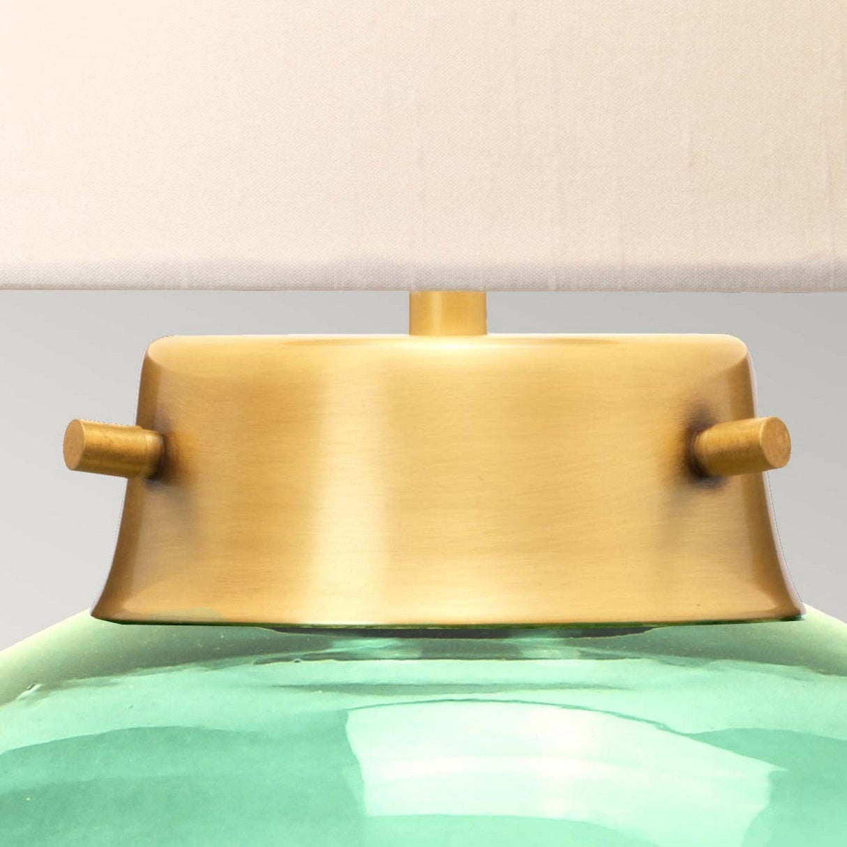 Close-up of the Kara Table Lamp - Aged Brass - Dark Green Glass, highlighting the aged brass base adorned with two small knob accents. The lower section is crafted from dark green glass and is complemented by a beige lampshade.
