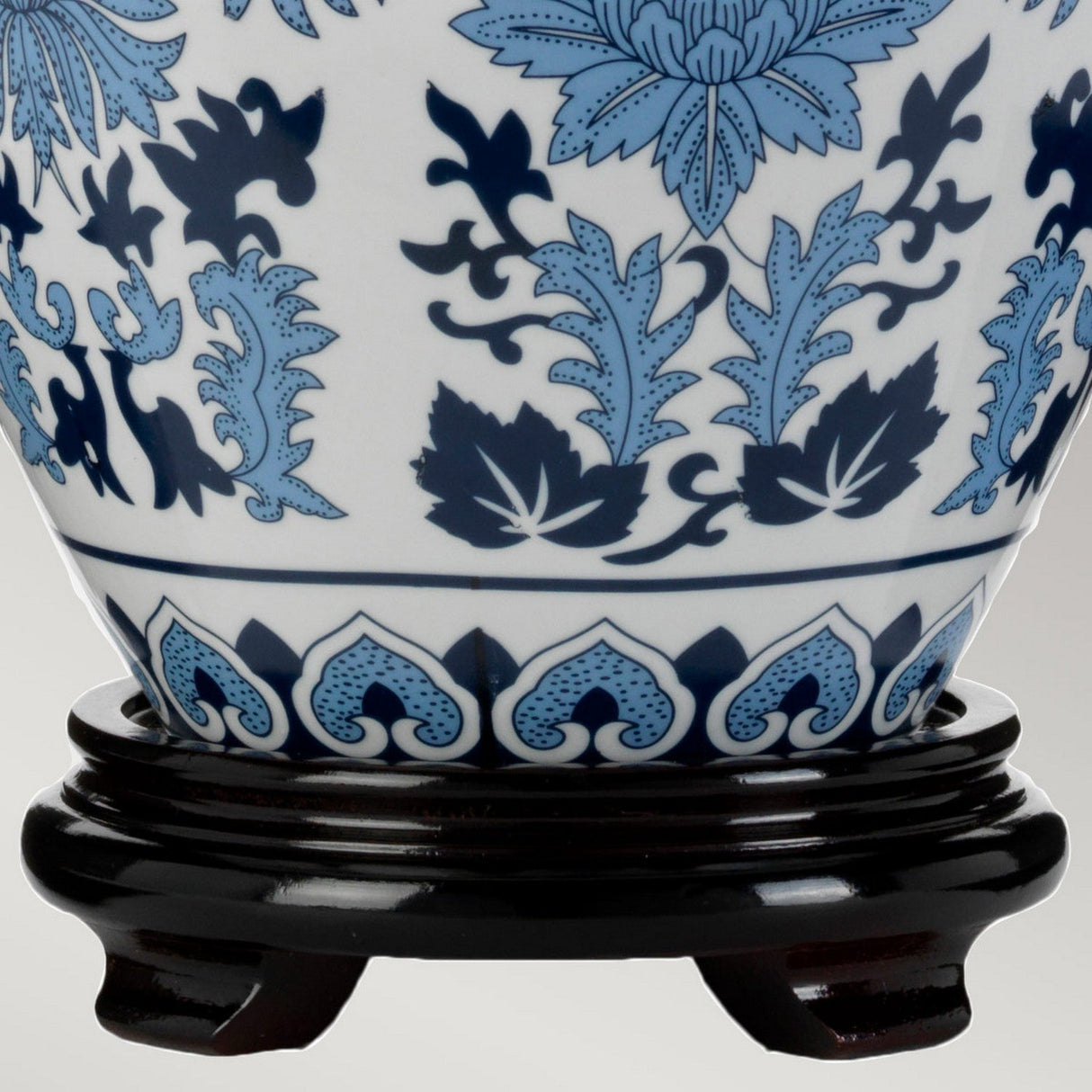 Close-up of a porcelain vase adorned with intricate blue floral and leaf patterns, reminiscent of the delicate designs on the Linyi Table Lamp With Tall Empire Shade - Blue & White. The vase rests on a dark wooden stand against a light gray background, highlighting its ceramic base.