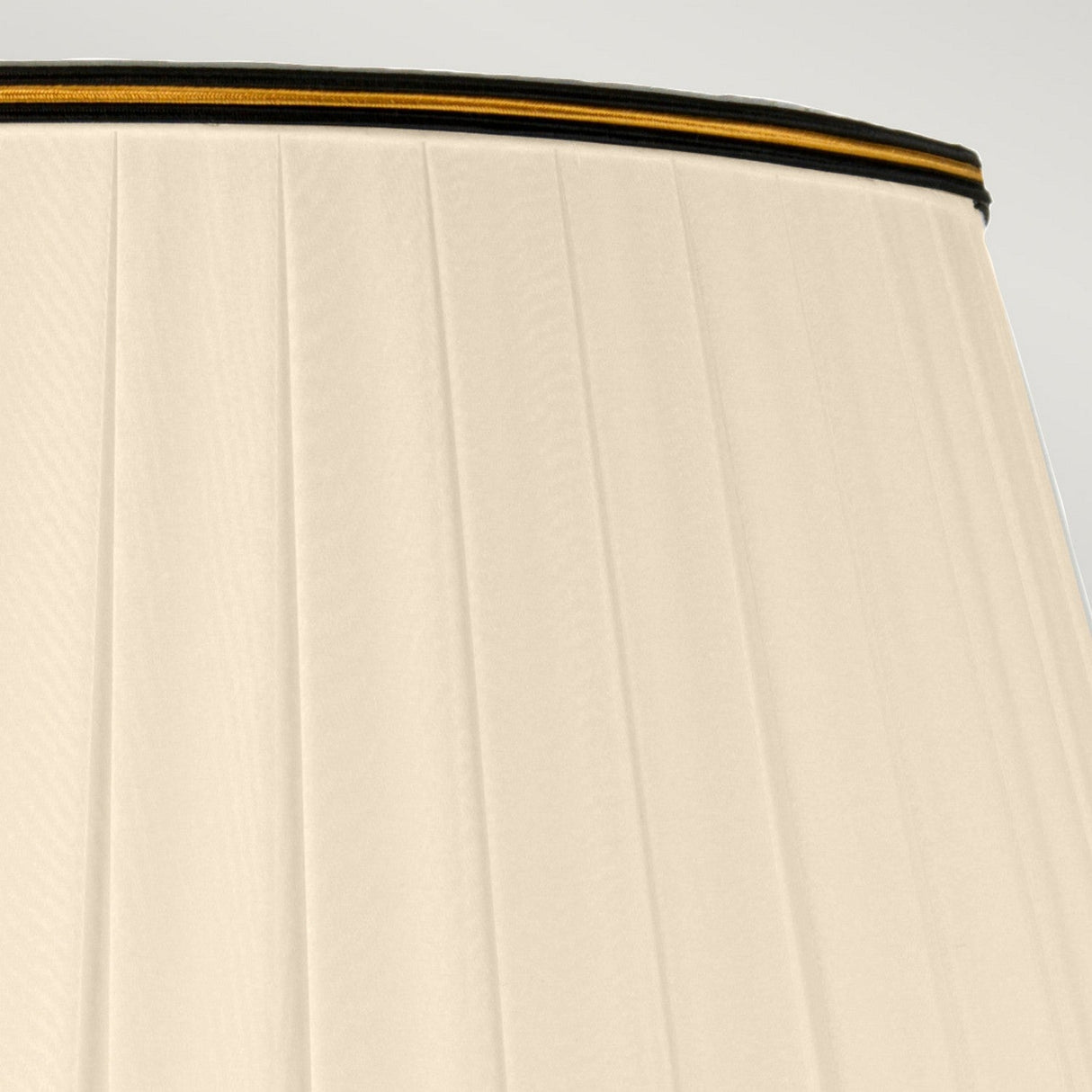 Close-up of a luxurious tall Empire shade in beige adorned with vertical pleats and accented by a black trim featuring a delicate yellow stripe at the top. The image beautifully showcases the fabric's texture and detail under soft lighting, perfectly complementing the Louviers Table Lamp in Oxblood & Gold.