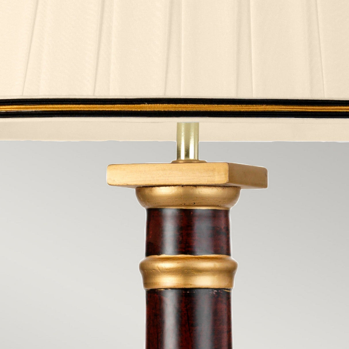 A close-up of the Louviers Table Lamp With Tall Empire Shade showcases its luxurious design, featuring a cream fabric shade with black and gold trim. The lamp's base is crafted from brass and dark wood, embellished with intricate gold accents. This elegant oxblood table lamp stands out against a plain light gray background.