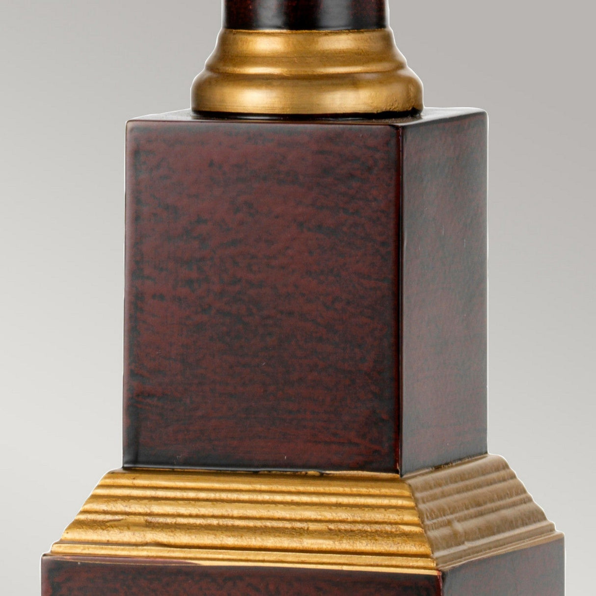 The decorative object, characterized by a square, wood-like base in dark brown and highlighted with gold accents, radiates luxury. It boasts a sleek, polished surface supporting its cylindrical top. This exquisite piece would beautifully complement the Louviers Table Lamp With Tall Empire Shade in Oxblood & Gold against a neutral gray backdrop.