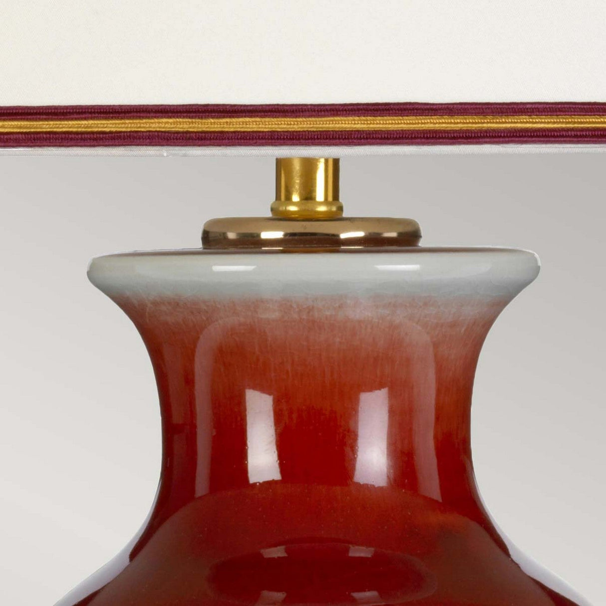 A close-up of the Majin Table Lamp With Tall Empire Shade - Oxblood, showcasing its glossy oxblood finish and a white fabric empire shade adorned with a thin red stripe near the top. The lamp's neck is accented with gold, while the lighting accentuates the smooth finish of its base.