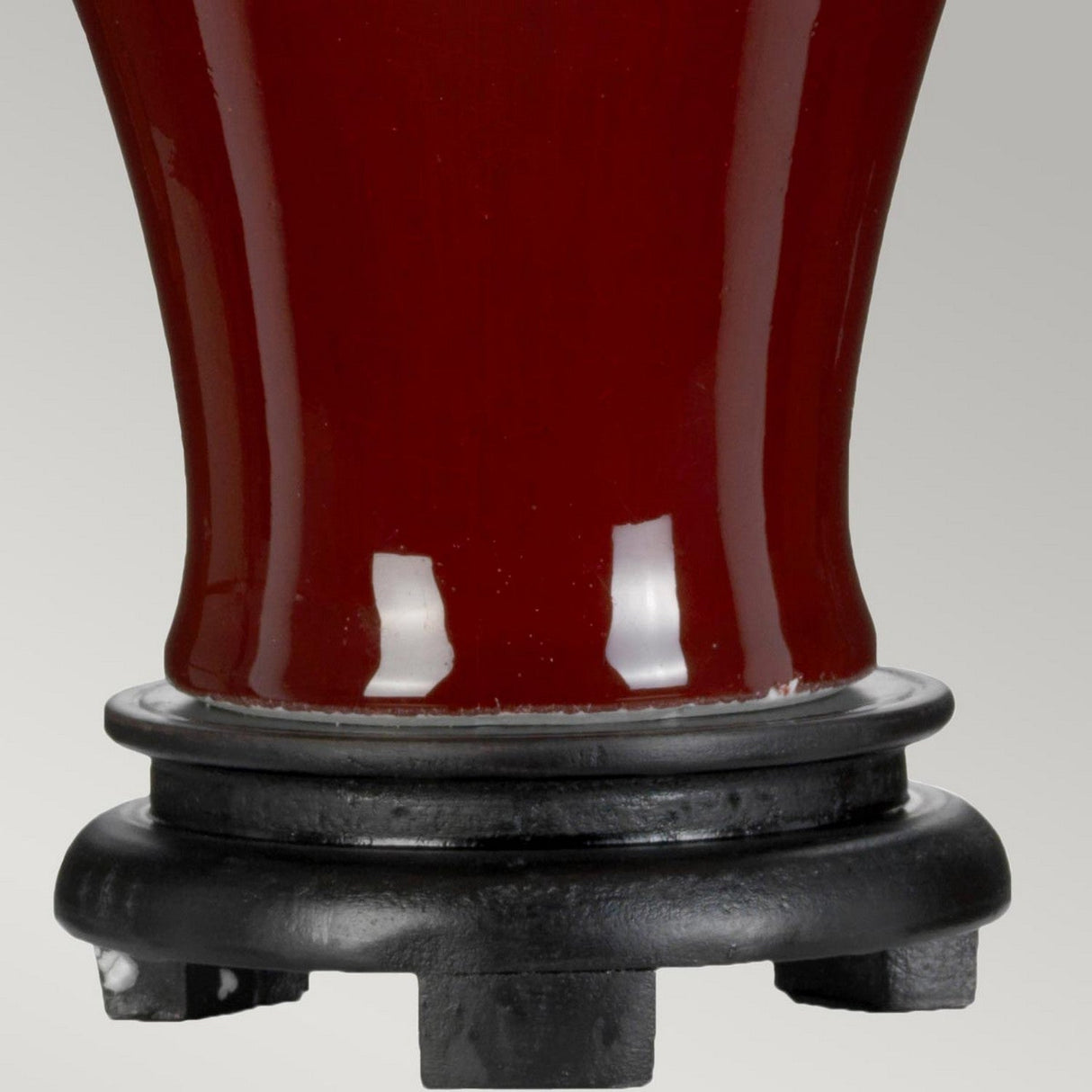 A close-up of the base of the Majin Table Lamp With Tall Empire Shade, featuring a sleek glossy red finish in oxblood, slightly flared in design, resting on a black circular base with intricately carved legs. The surface beautifully reflects light, accentuating its shiny finish.