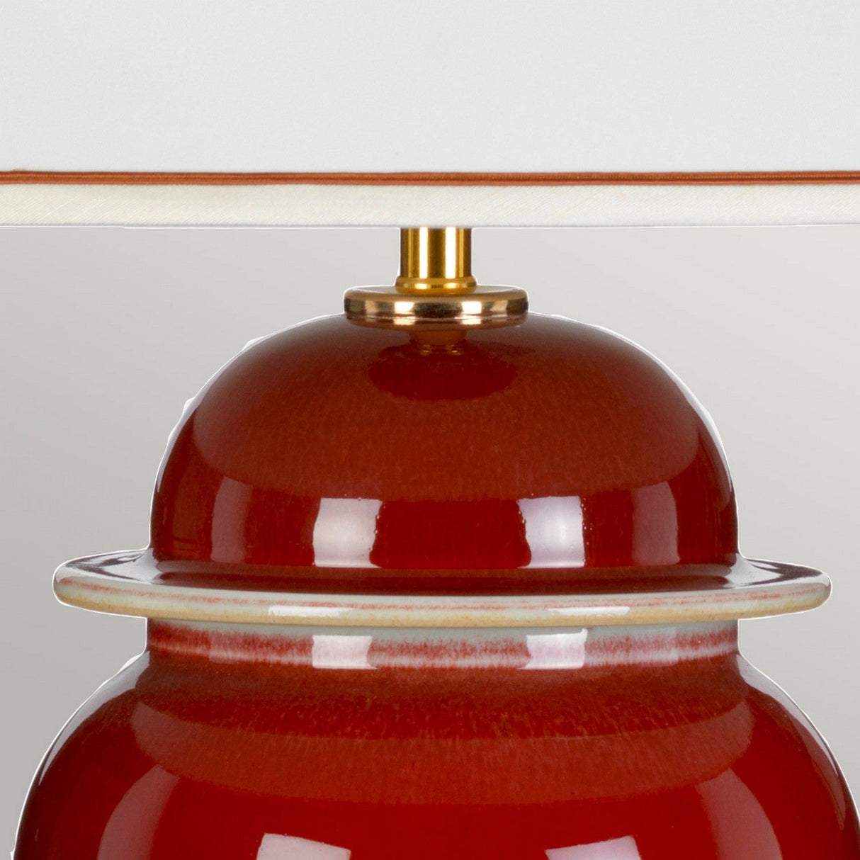 Close-up of the vibrant Oxblood Temple Jar Table Lamp With Tall Empire Shade - Oxblood, showcasing its glossy finish. The lamp is paired with a beige lampshade and topped with a brass element, exuding oriental elegance that is brought to life by soft lighting.