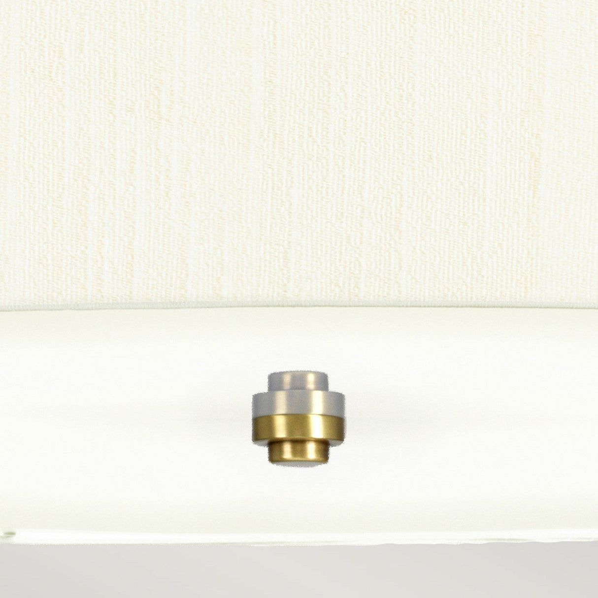 A metallic-colored, cylindrical knob resembling the Pearce 30" Pendant Light in aged brass, with an ivory-toned finish, is mounted on a textured white wall panel, centered just above a smooth ledge.