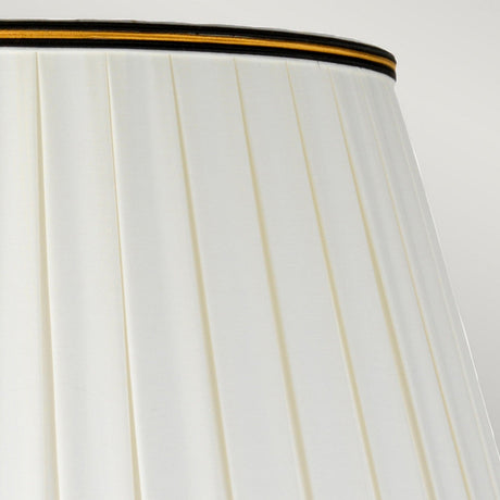 A close-up of the finely textured Peronne Table Lamp with its tall Empire shade showcases a pleated white design adorned with black and gold trim near the top edge, set elegantly against a neutral background.