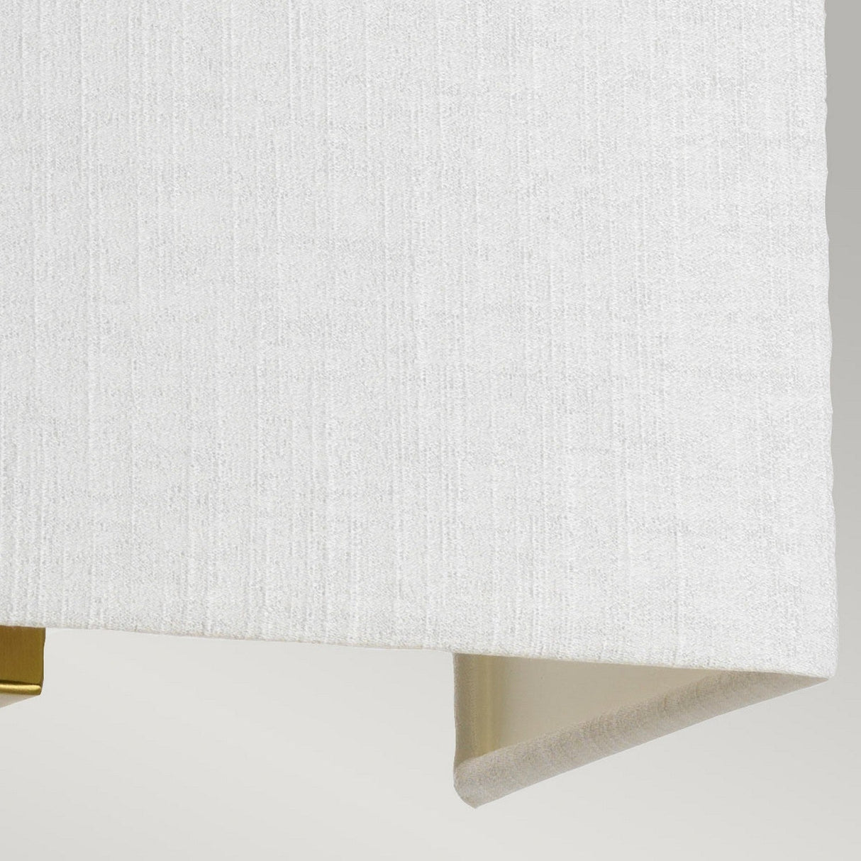 A close-up view of the Riley Medium Rectangular Wall Light in Aged Brass highlights its beige, textured fabric with a subtle woven pattern that evokes the look of a faux silk shade. The material rests against a light gray background, elegantly folding at the edges to reveal its varied tones underneath.