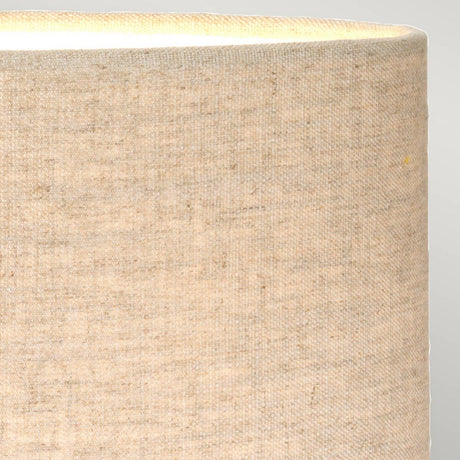 Close-up of a beige fabric lampshade with a soft texture on the Sabi Table Lamp With Oval Shade - Bronze Patina. Its top edge emits a subtle glow, indicating it's illuminated. The shade's neutral hue beautifully complements the bronze patina finish, enhancing its minimalist design.