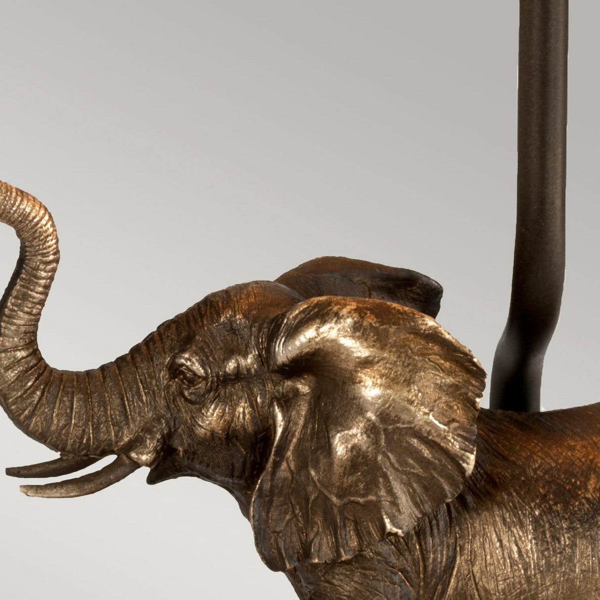 The Sabi Table Lamp With Oval Shade - Bronze Patina features an exquisite bronze elephant sculpture with a rich patina finish, showcasing detailed elements such as a raised trunk and textured ears. Against a plain gray background, it serves as the majestic base for this elegant lamp.