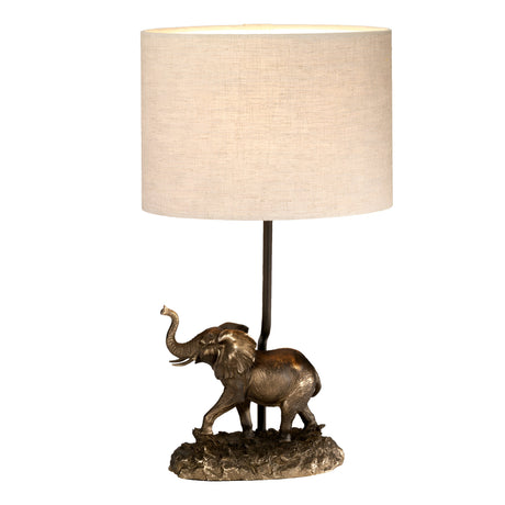 The Sabi Table Lamp With Oval Shade - Bronze Patina highlights a decorative base with a bronze elephant statuette, its trunk raised, complemented by an elegant beige cylindrical lampshade.