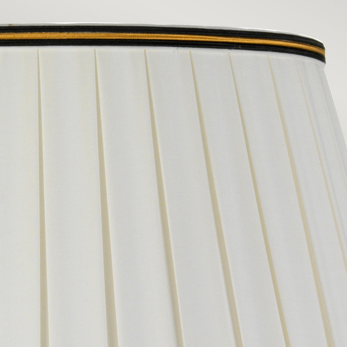 Close-up of the top section of a Sashi Table Lamp with Tall Empire Shade, highlighting its green pleated fabric with oriental craftsmanship. A gold and brown band trims the top edge, enhancing its elegance. The ambient light casts a gentle glow against the plain, light gray background.