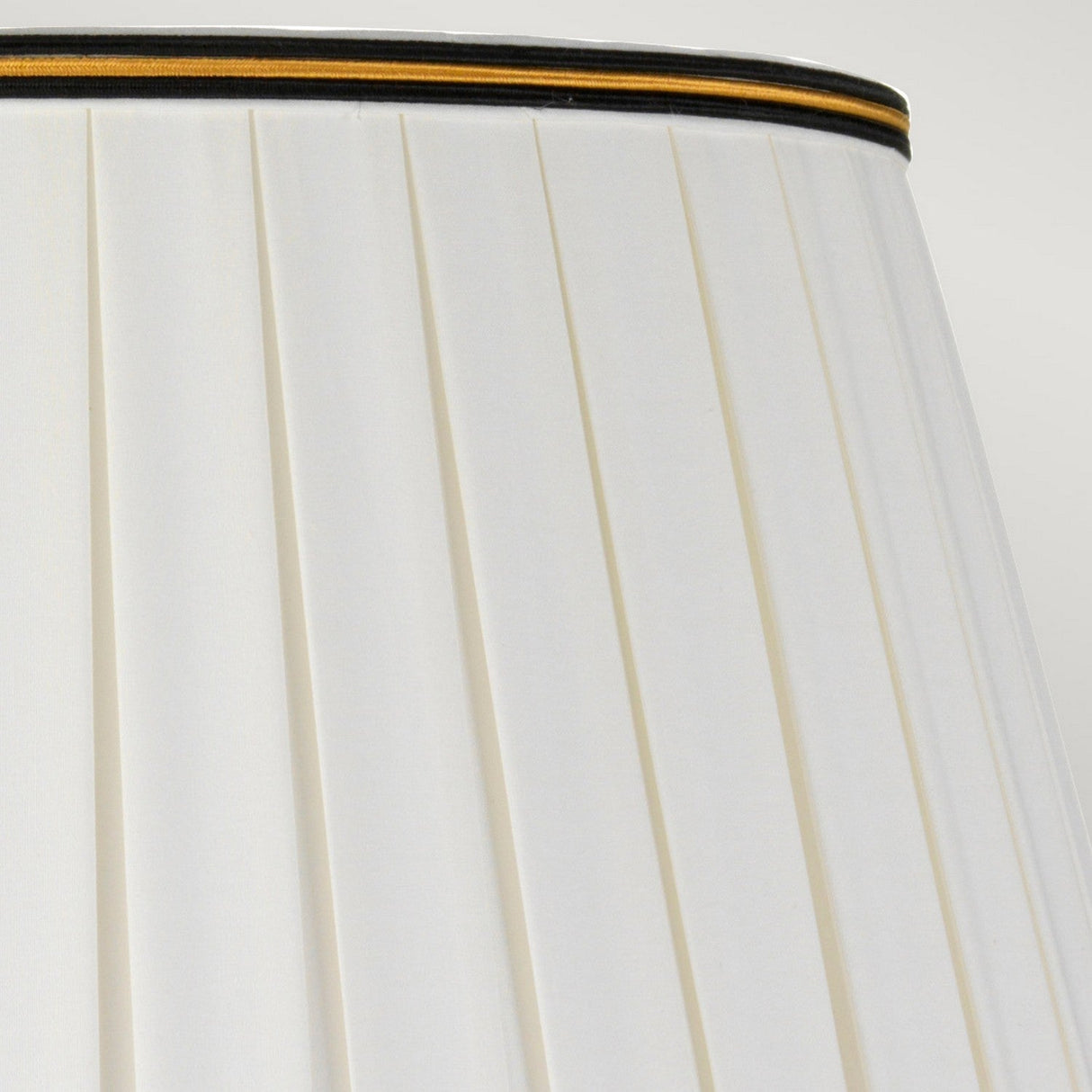 Close-up of a pleated white lampshade on the Taipei Table Lamp With Tall Empire Shade - Black & Gold, showcasing a slender black and gold trim along the top edge. The light gray background beautifully complements this exquisite piece of black and gold lighting.