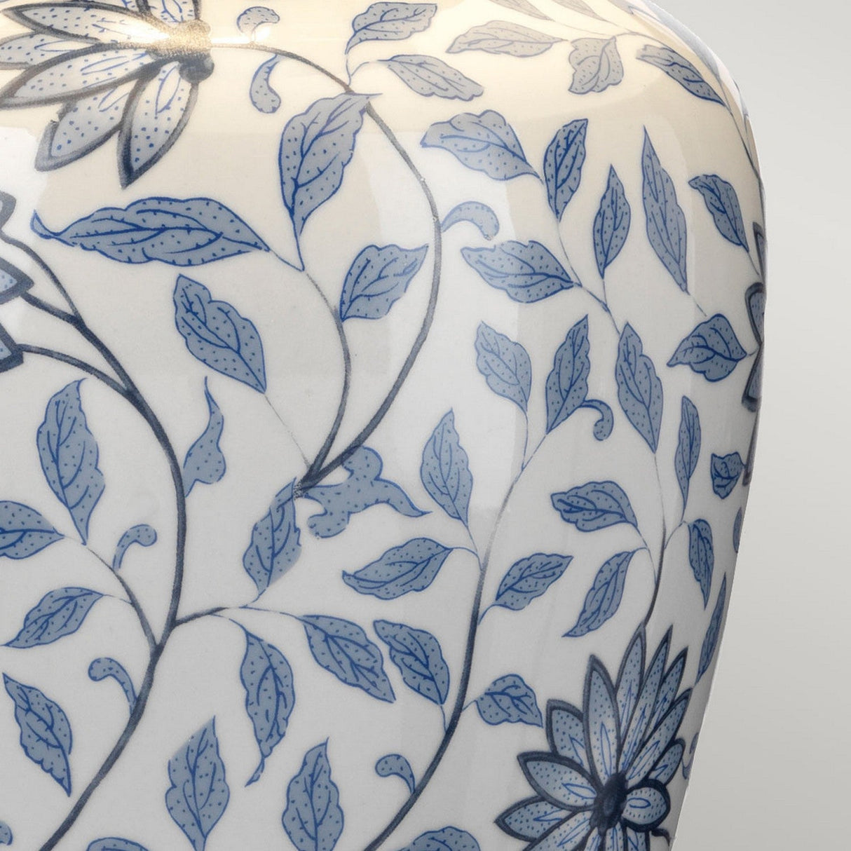 A close-up of the Ying 1 Light Table Lamp showcases its blue and white porcelain base adorned with a floral pattern reminiscent of classic Chinese designs. The intricate leaves and flowers are highlighted on a white backdrop, all beautifully illuminated by soft ambient lighting, offering a timeless artistic appeal.