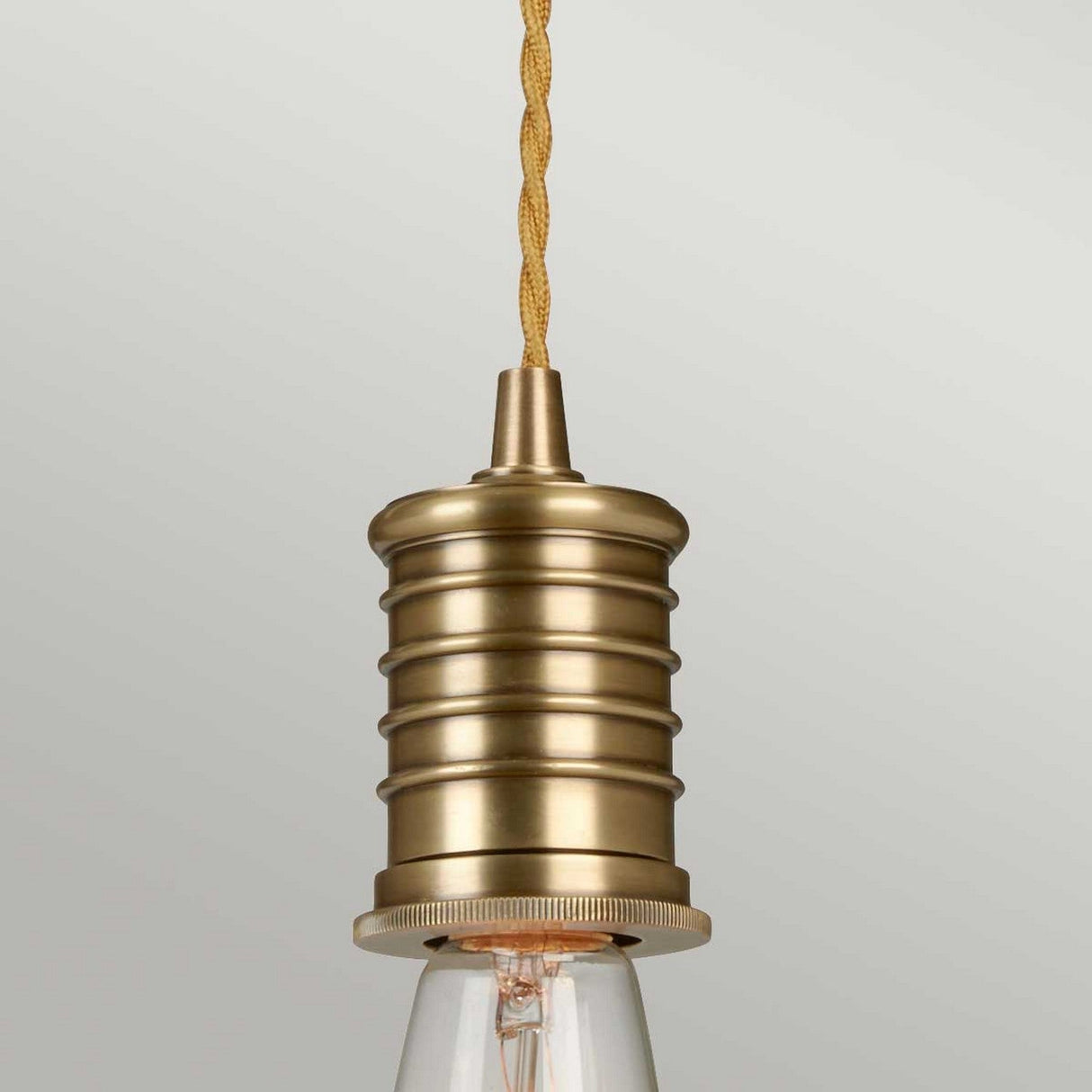 The Douille 1 Light Pendant in Aged Brass showcases a vintage design with ribbed detailing and a twisted fabric cord. Its exposed LED filament lamp adds to its retro charm, seamlessly merging classic and modern styles.