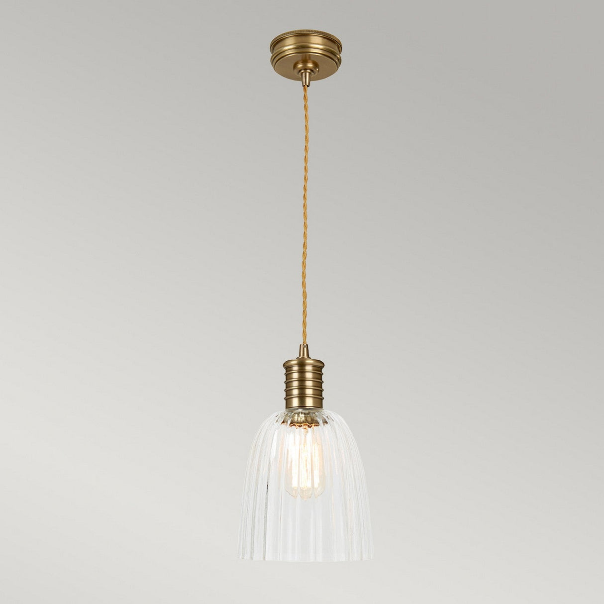 The Douille 1 Light Pendant in aged brass features a vintage design with a twisted cord and ribbed, translucent glass shade that hangs from the ceiling. It casts a warm glow against neutral surroundings, enhanced by an LED filament lamp.