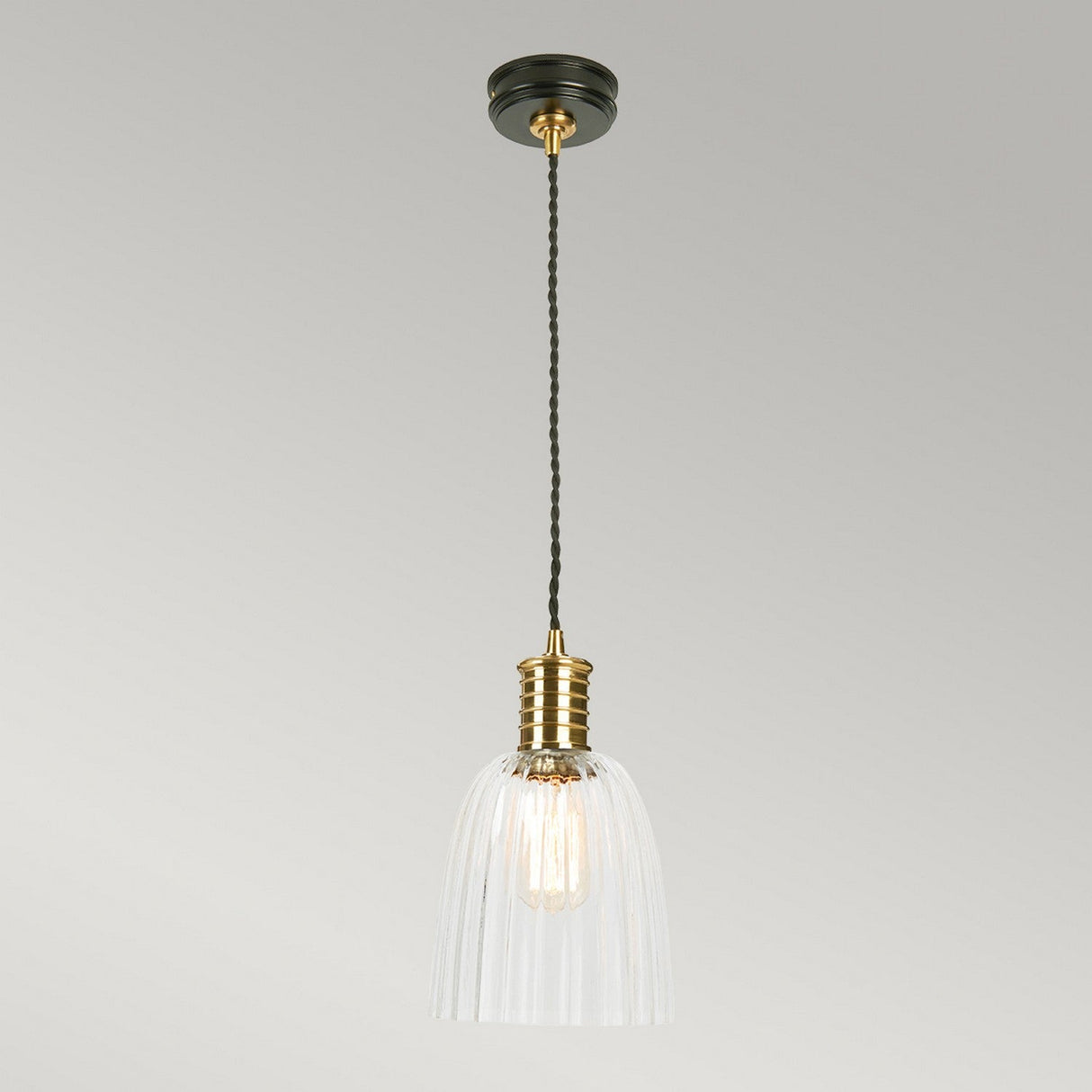 The Douille 1 Light Pendant, featuring a polished brass lamp holder, a clear ribbed glass shade, and an industrial style with a twisted black cord, hangs from the ceiling against a gray background.