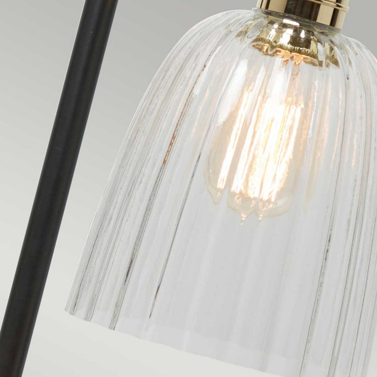 A close-up view of the Douille Table Lamp - Black & Polished Brass reveals its lit clear glass with a ribbed design, highlighting the glowing bulb within. The lamp boasts a sleek black metal frame complemented by polished brass accents, merging modern elegance with a touch of vintage style.