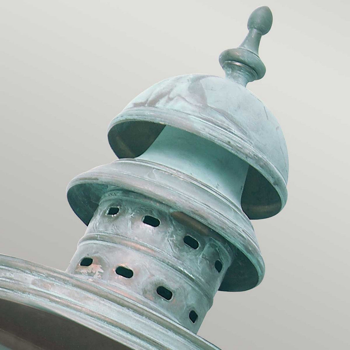A close-up of the Downing Outdoor Wall Light in Turquoise reveals its weathered finish, evocative of a pre-Georgian lantern, with a domed top and decorative finial. The middle section is adorned with round perforations, all set against a soft, muted background.