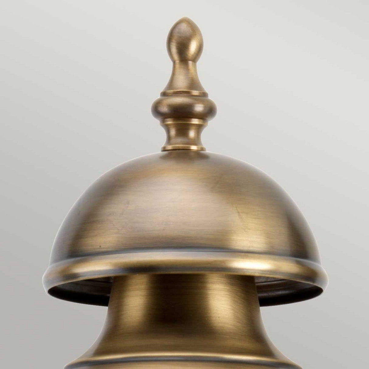 A close-up of the Downing Outdoor Wall Light in aged brass showcases its metallic, antique-finish decorative finial with a dome-shaped base and pointed top against a light gray background. The design highlights smooth curves and a shiny texture reminiscent of an elegant exterior decor piece.