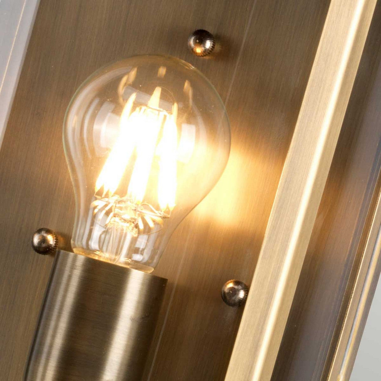 A close-up of the Downing Outdoor Wall Light in aged brass shows a glowing bulb that emits a warm, yellowish light, enhancing the polished surface of the metal. The light fixture is mounted with visible screws, adding a touch of vintage charm to its design.