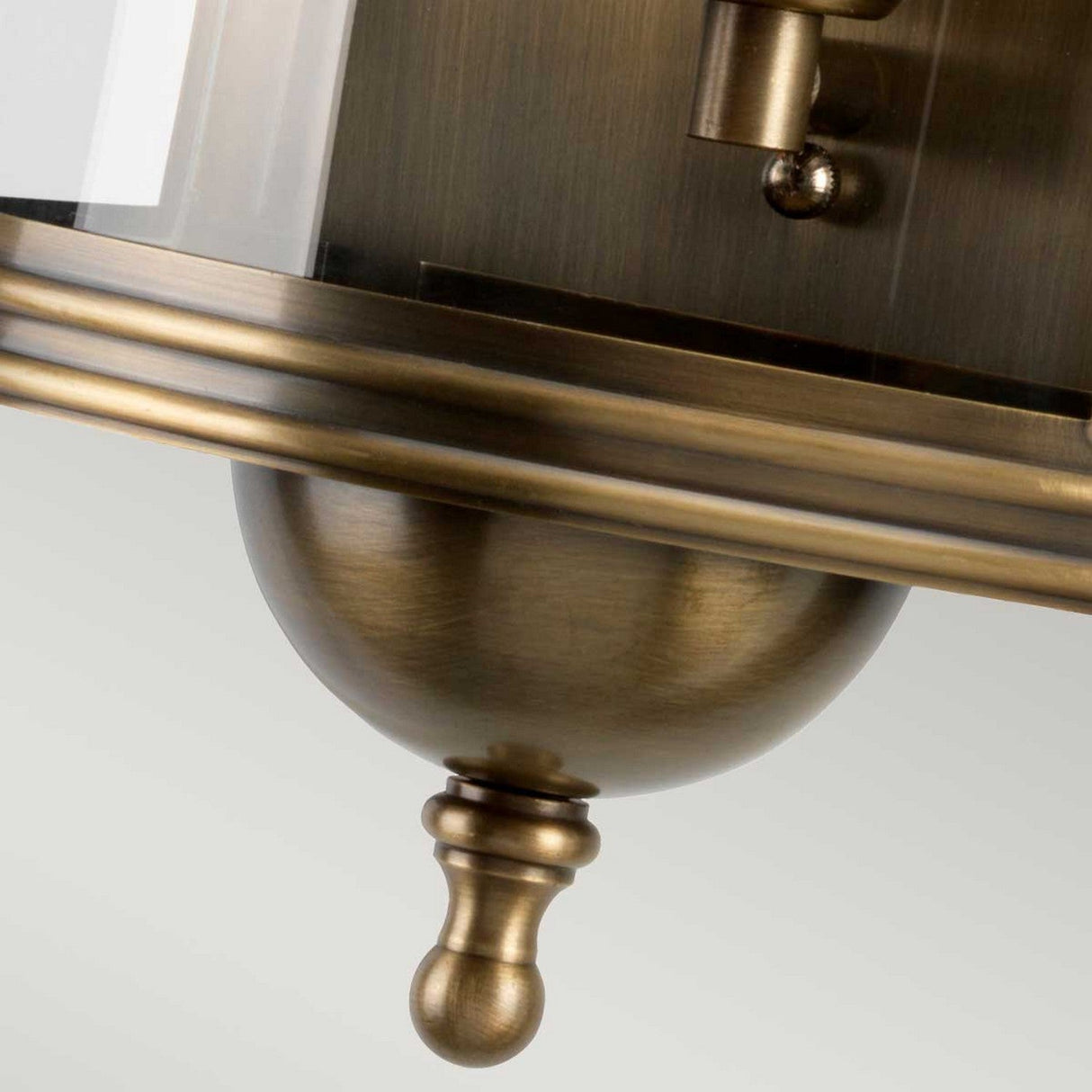 Close-up of the Downing Outdoor Wall Light in an aged brass finish, showcasing its vintage-style charm. It features a rounded base and decorative knob with part of the lamp shade visible in the background. This fixture boasts an elegant, classic design perfect for enhancing any exterior space.