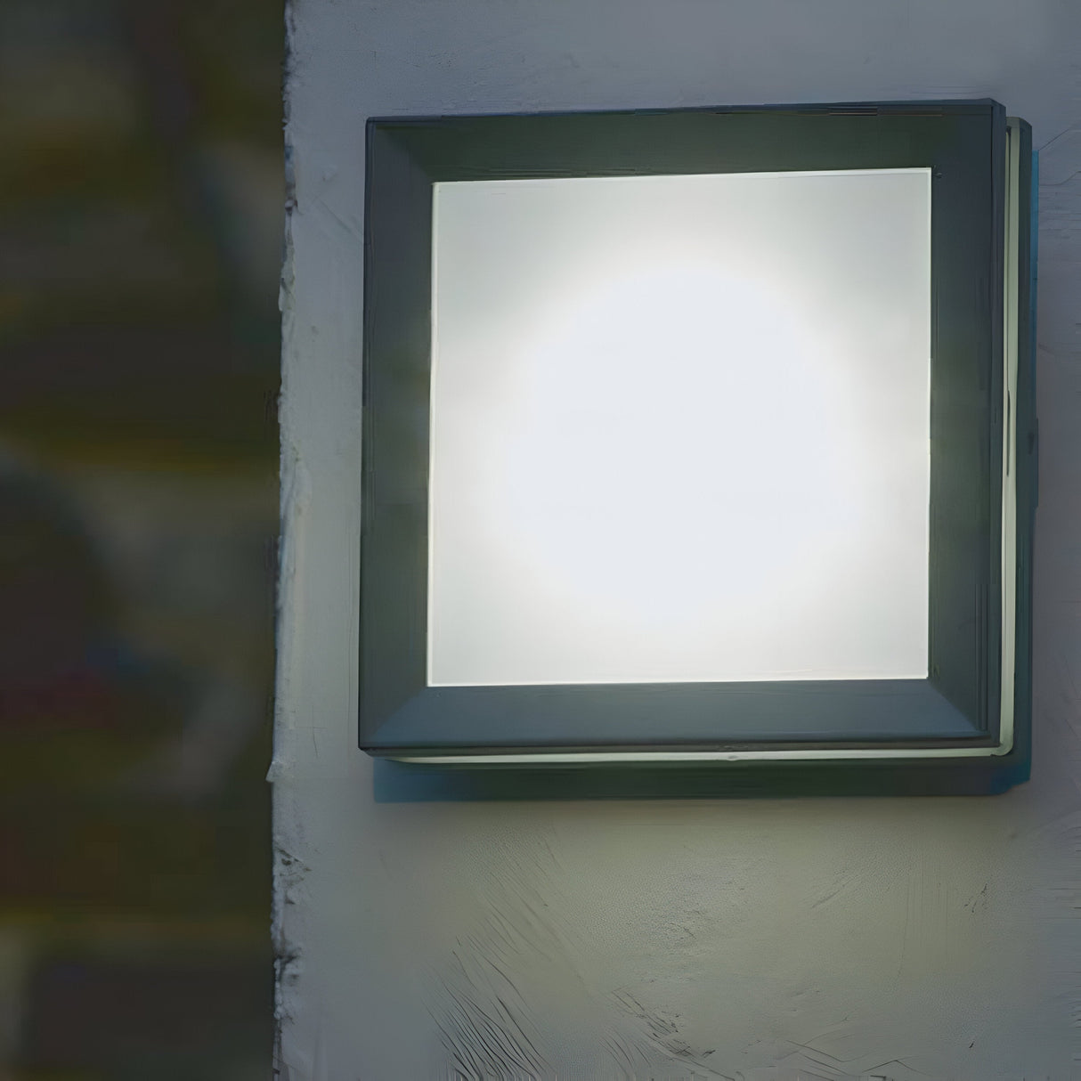 The Egil-1 Outdoor Wall/Ceiling Light in graphite casts a bright, glowing white light against a light gray wall. Made from cast aluminum with a sleek graphite finish, the fixture features a minimalist and modern design highlighted by its dark frame encircling the illuminated area.