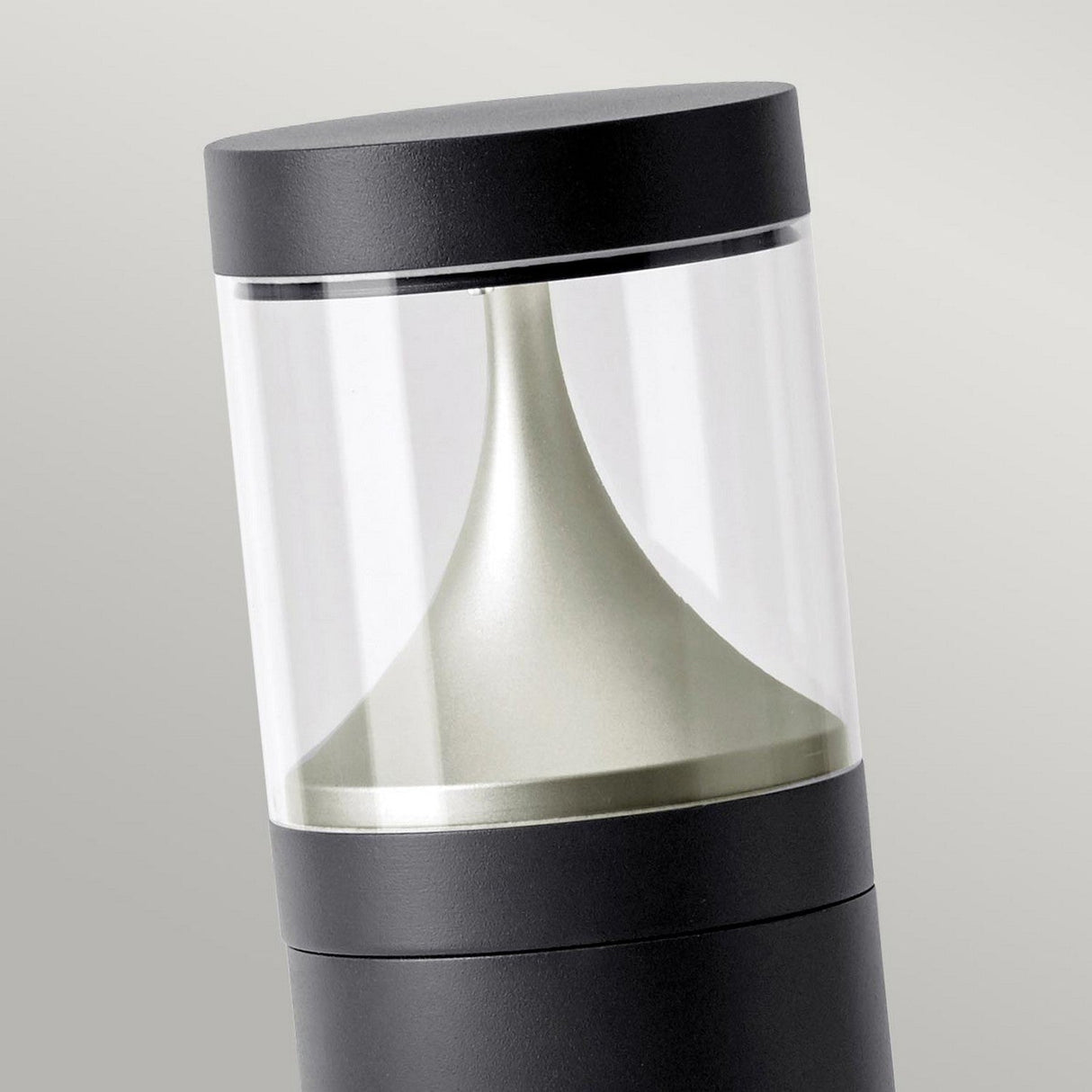 The Egersund LED Tall Bollard - Graphite showcases a sleek, contemporary design with a black and transparent cylindrical structure complemented by an internal metallic conical component, all set against a light gray backdrop. This modern outdoor lighting solution utilizes energy-efficient LED technology to provide sustainable illumination.