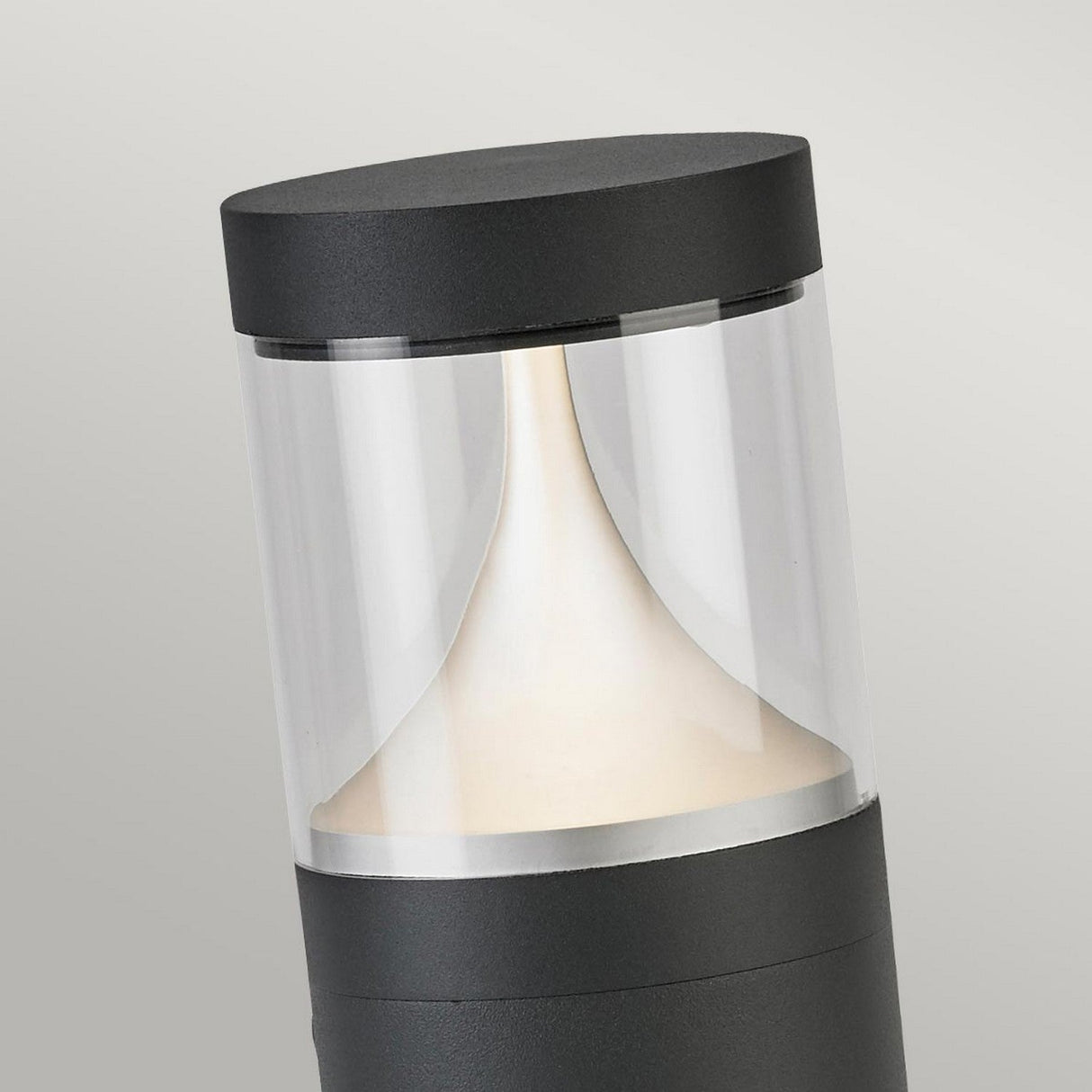 The Egersund LED Short Bollard in Graphite boasts a contemporary cylindrical design with a clear glass segment and a sleek dark gray finish. Offering energy-efficient lighting, it emits a soft, warm glow that elegantly highlights the curved interior surface with its welcoming charm.