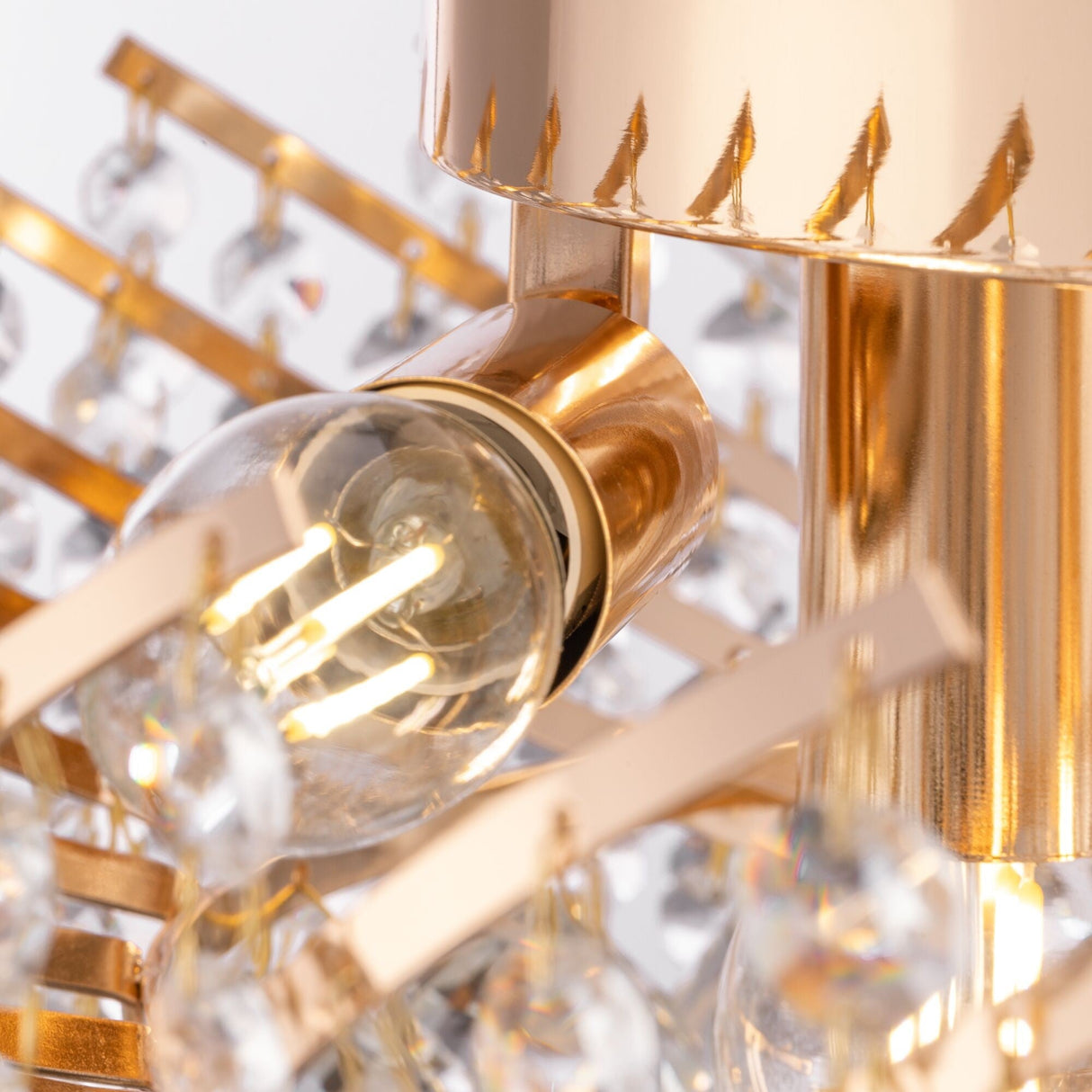The Karolina 6 Light Chandelier - Gold showcases a stylish design with a clear light bulb, visible filaments, gold metal accents, and dangling crystal pendants, offering a luxurious and sophisticated look ideal for classic interiors.