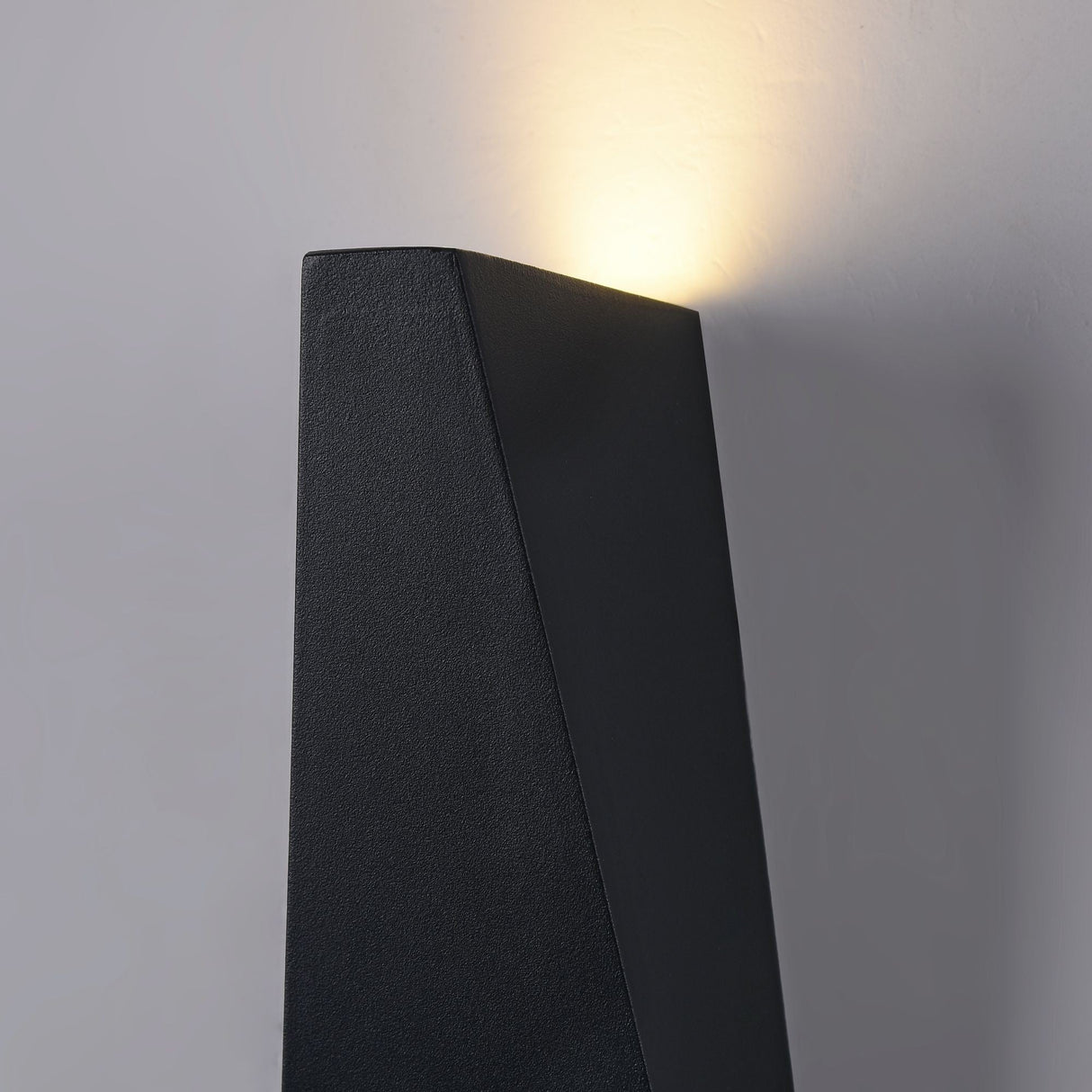 The Times Square Outdoor Wall Light 3200K 6W - Black, made from corrosion-resistant aluminium, showcases a sleek geometric design that emits a soft, warm glow against the light gray wall, enhancing a minimalist and contemporary aesthetic.
